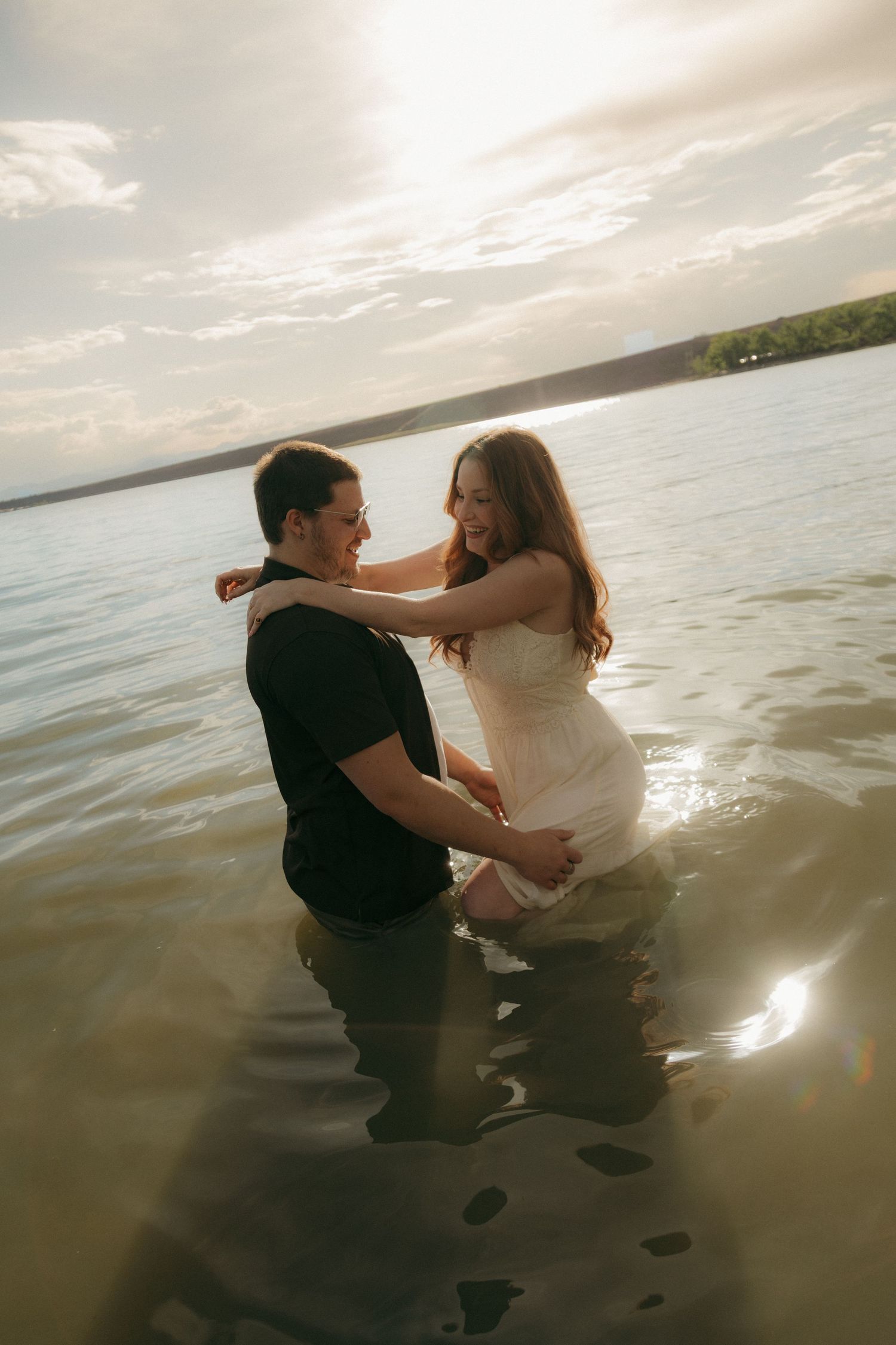 Couple sitting in sunlit lake water, woman in white dress smiling at man in black shirt.