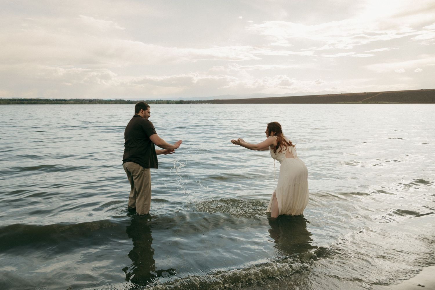 Couple playfully splashing each other while wading in a calm lake during golden hour engagement photos.