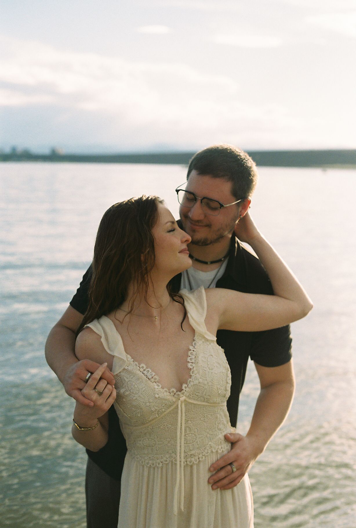 Couple sharing a tender moment by a lake at sunset, woman in lace dress with man in black shirt.