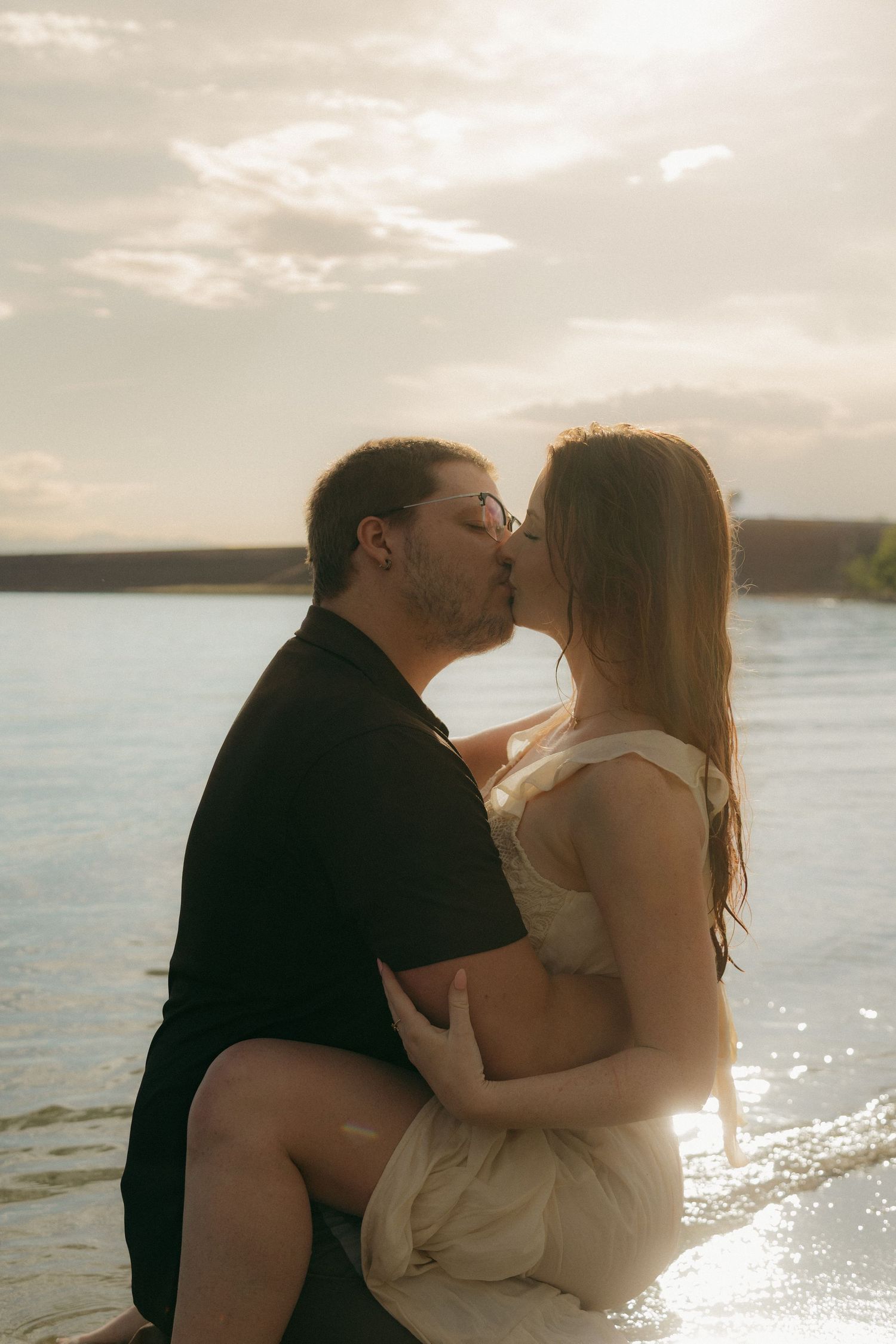 Couple sharing a tender kiss while sitting on beach shore with golden sunset light behind them.