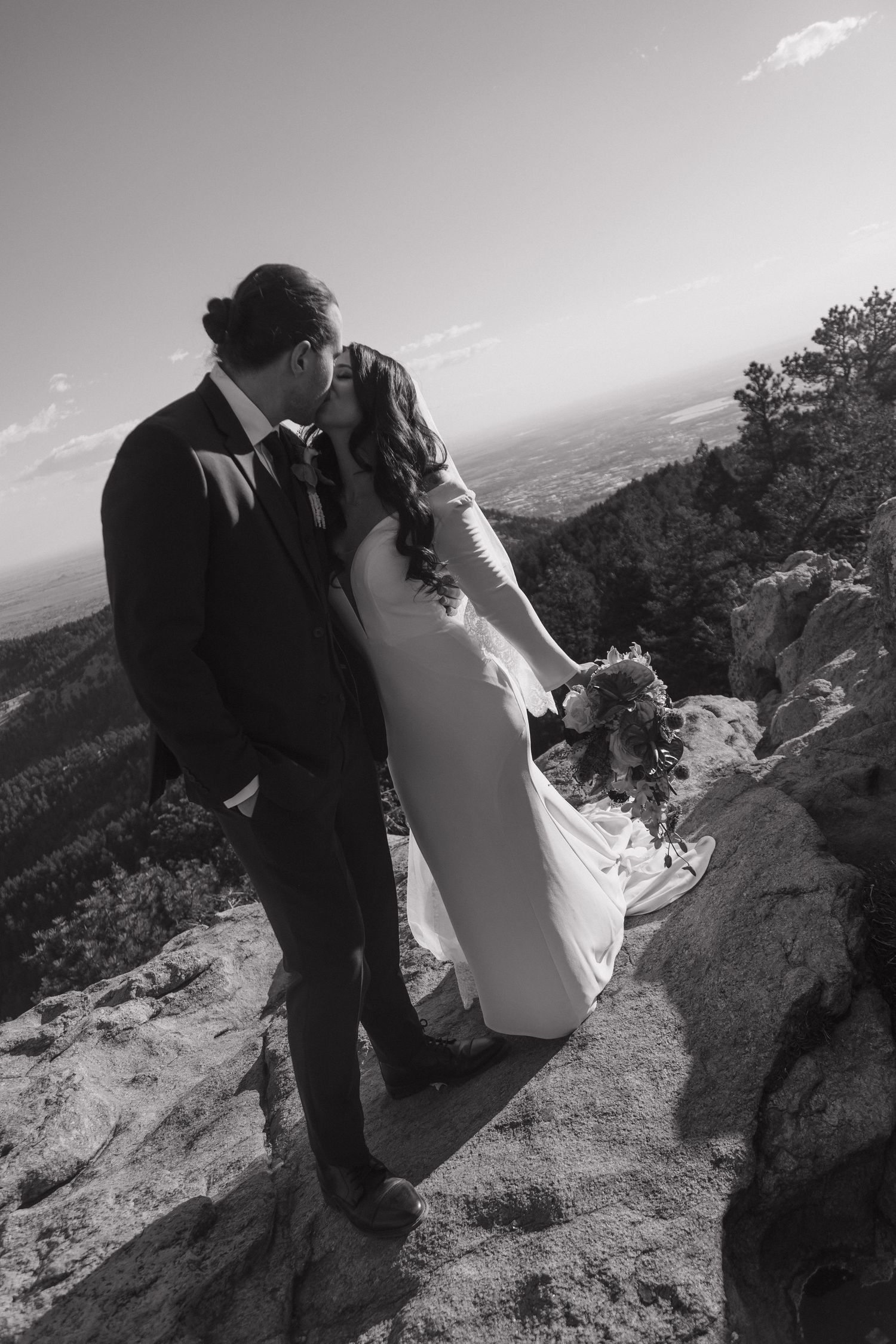 Bride and groom kissing on rocky mountain summit with panoramic landscape in black and white.
