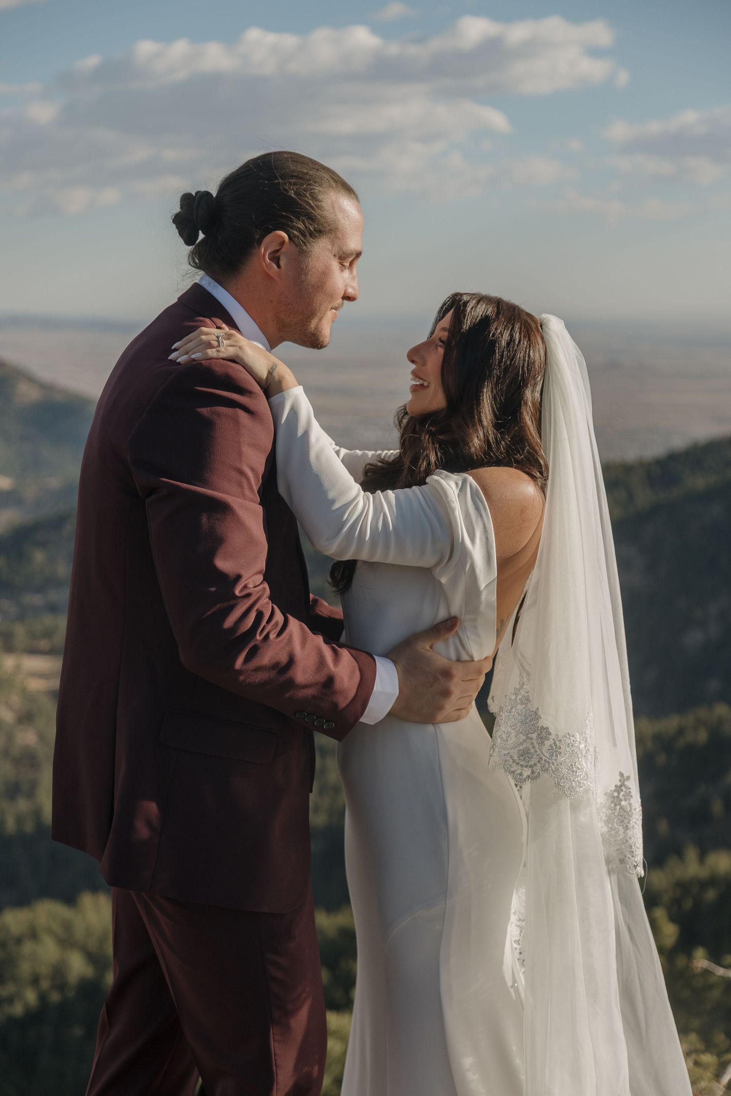 Bride in white gown and groom in burgundy suit embrace and smile at mountain elopement ceremony.