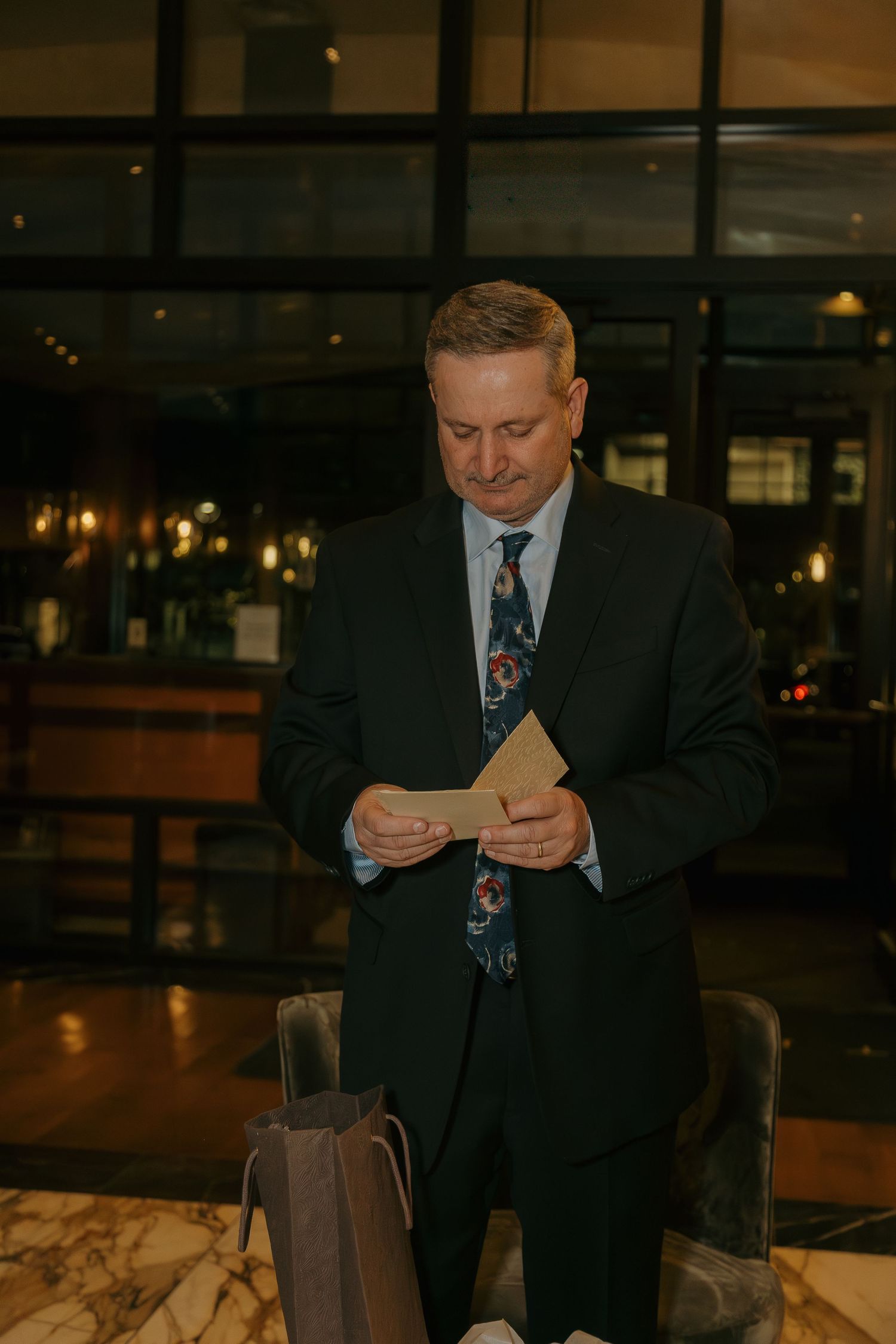 Dapper man in floral tie reads a wedding card while standing in a warmly lit hotel lobby.