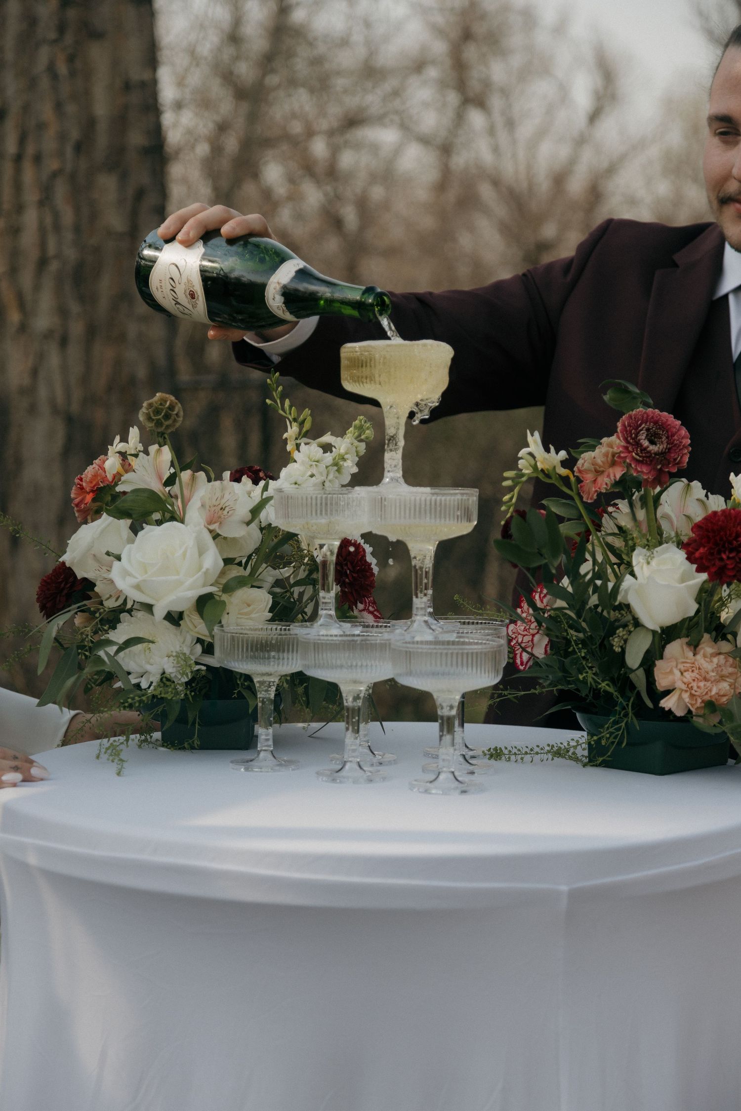 Groom pouring champagne tower at elegant wedding reception table adorned with white and burgundy florals.