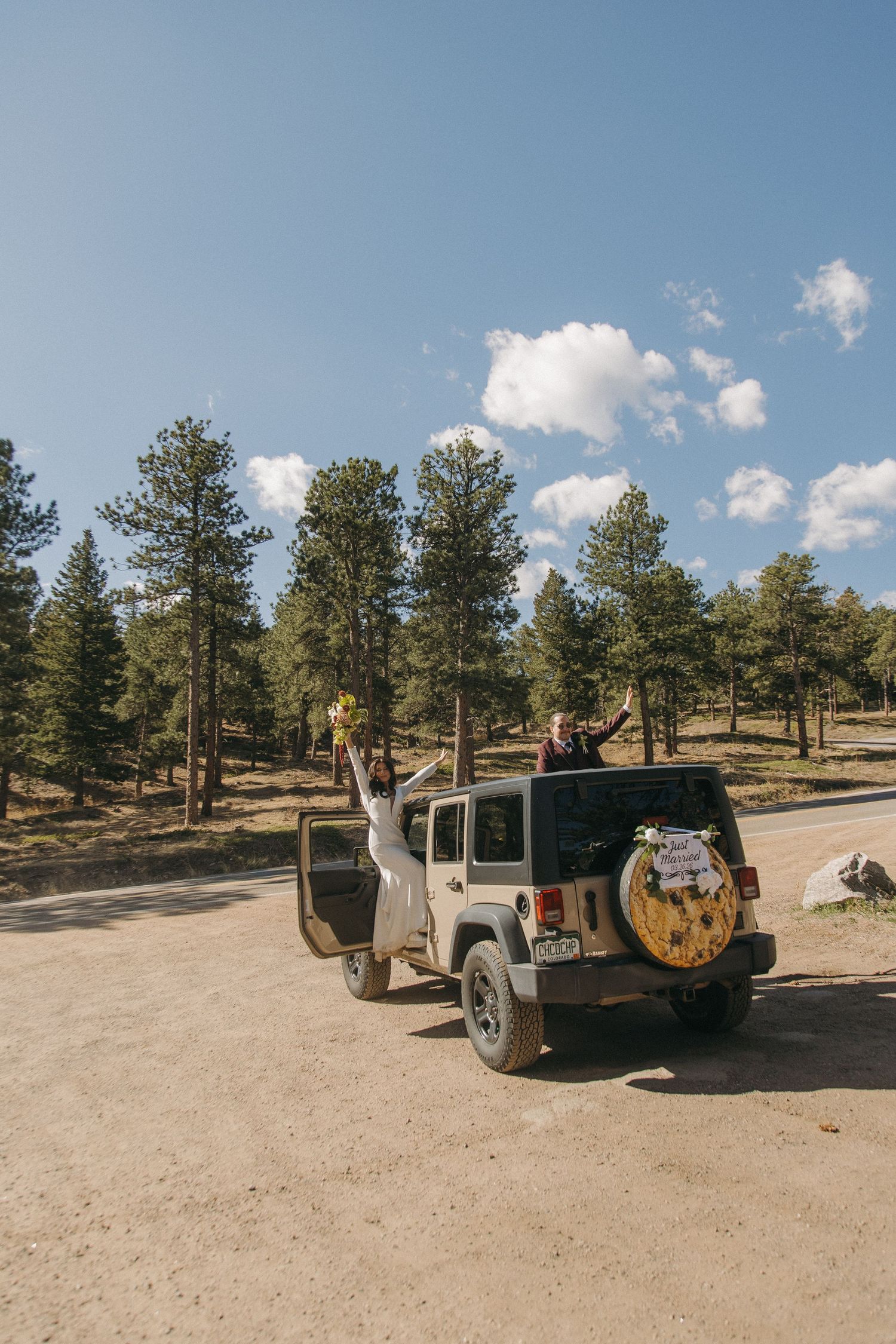 Joyful bride and groom celebrate beside decorated Jeep on forest road under blue sky.