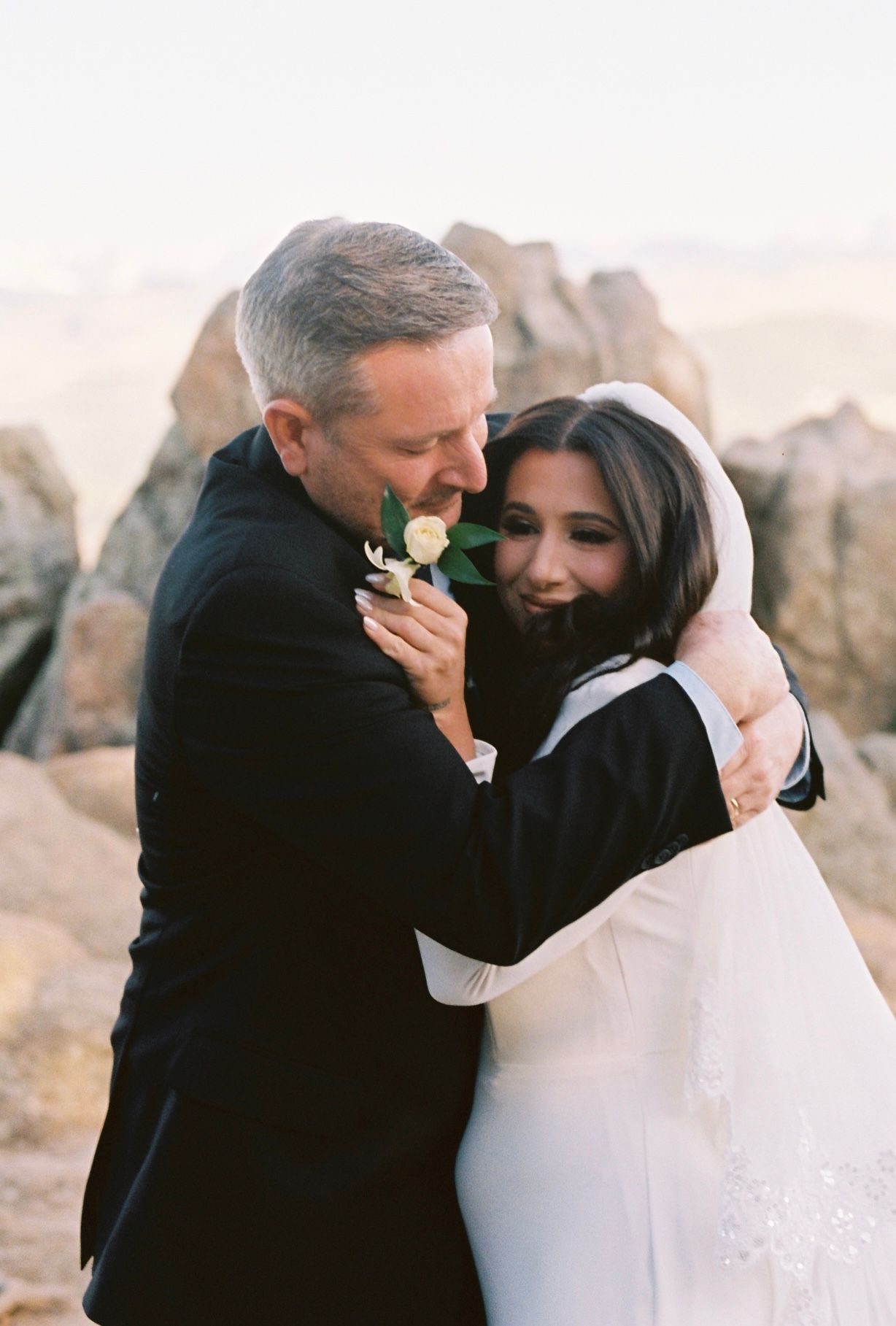 Groom in navy suit embracing smiling bride in white veil against rocky desert landscape backdrop.