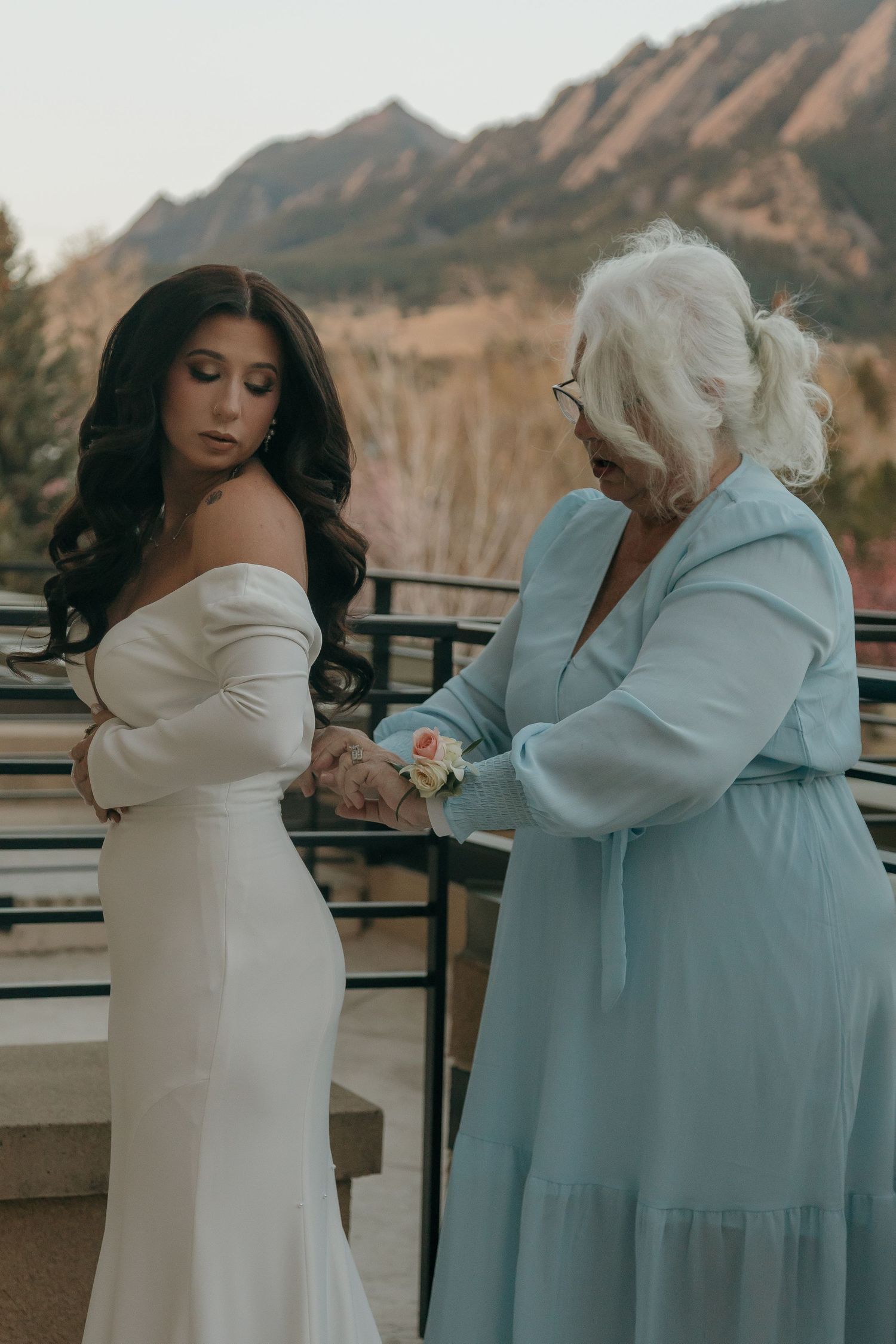 Older woman in blue dress pins corsage on bride in white off-shoulder gown outdoors.
