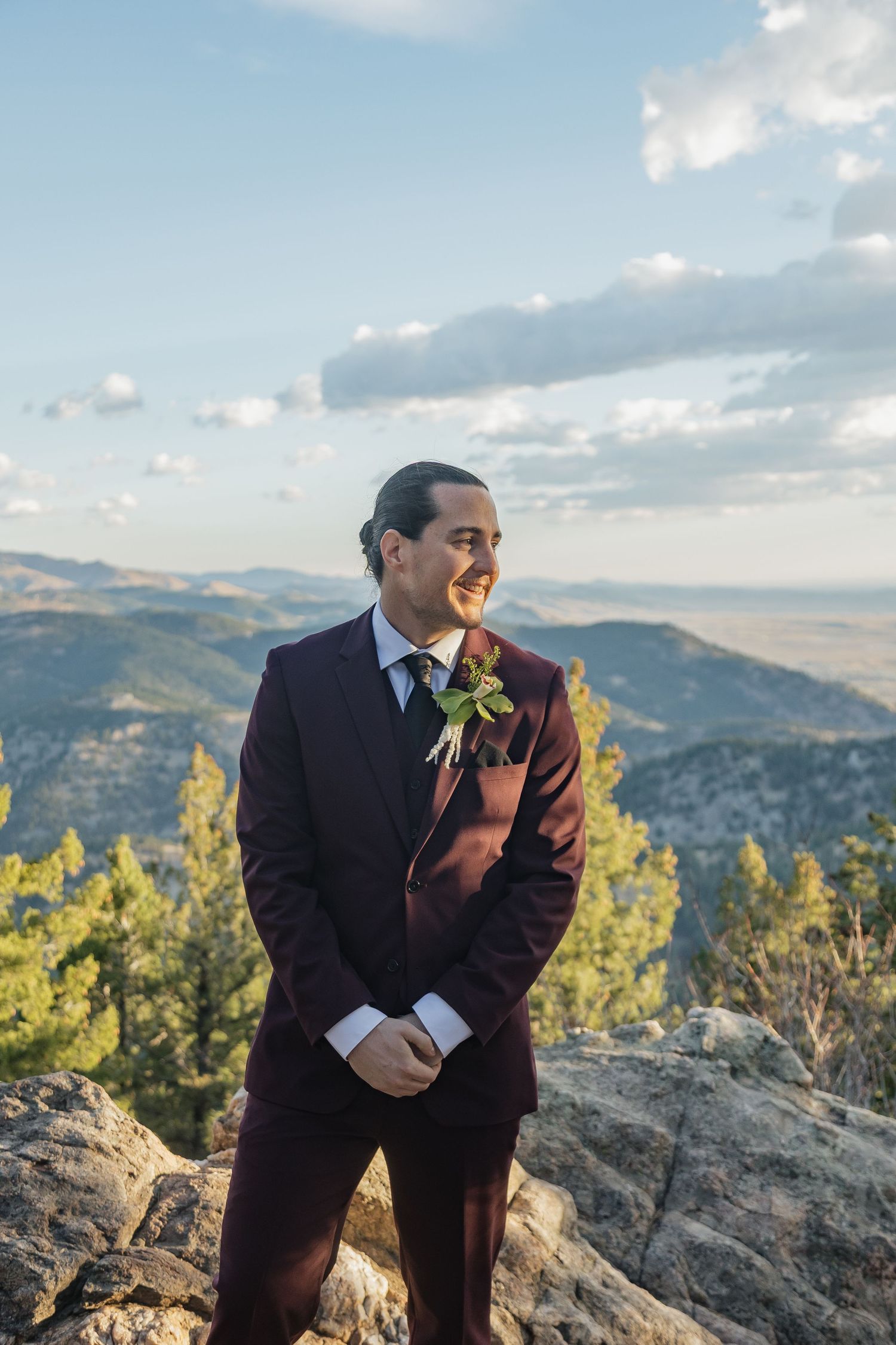 Handsome groom in burgundy suit with boutonniere smiling on rocky mountain summit with scenic valley views.