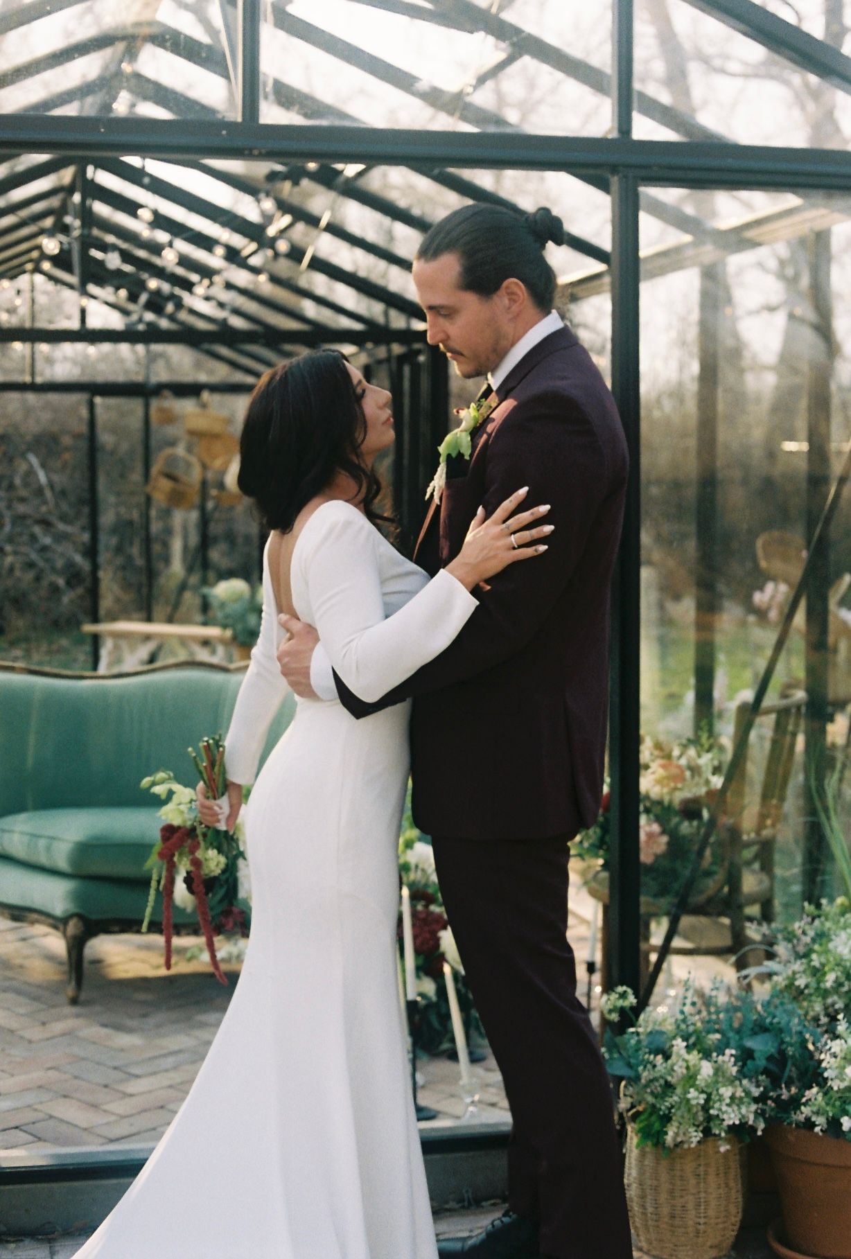 Bride and groom share intimate moment in front of glass greenhouse surrounded by plants.