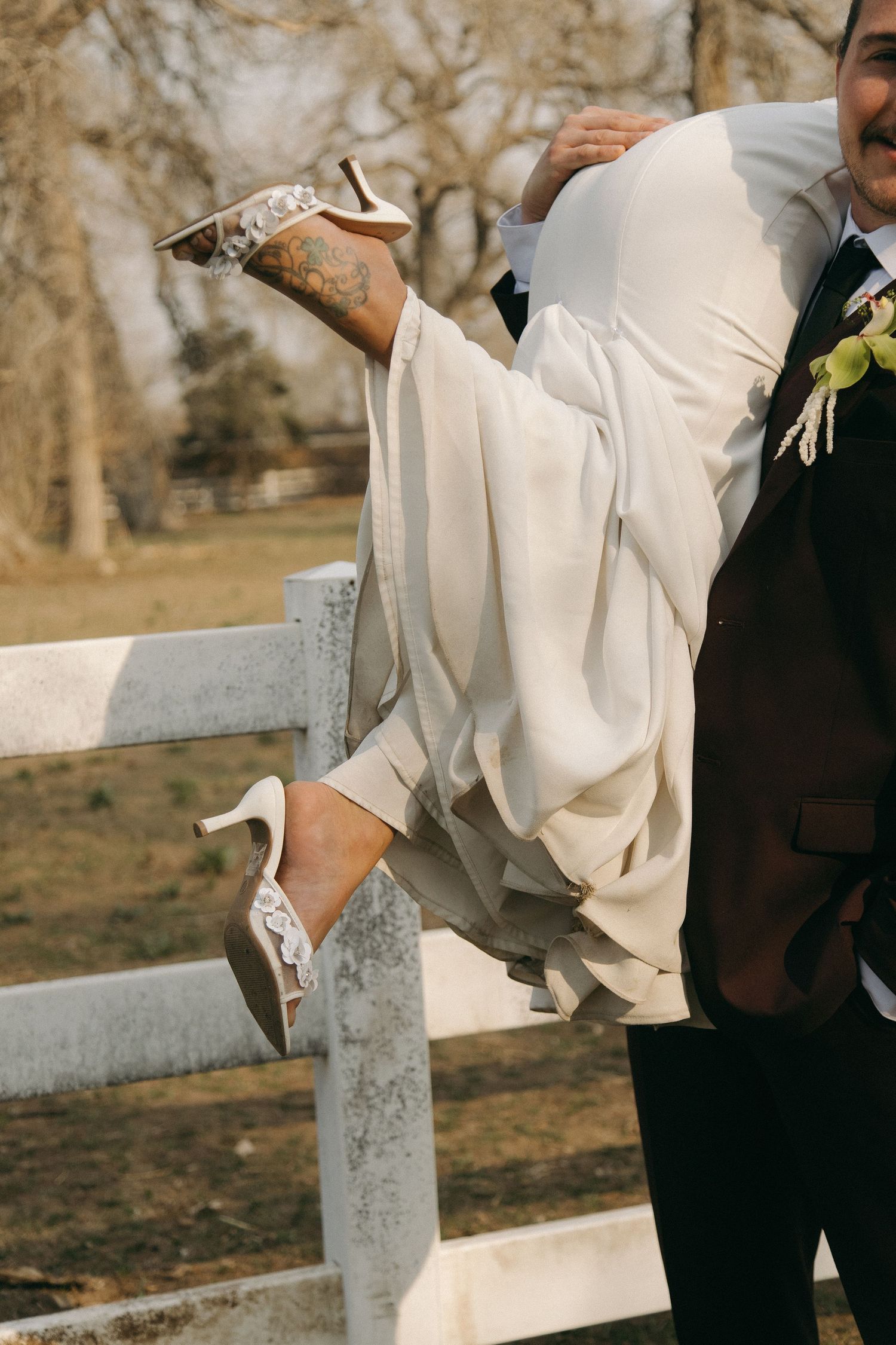 Groom in dark suit carrying bride in white gown near white fence at outdoor wedding.