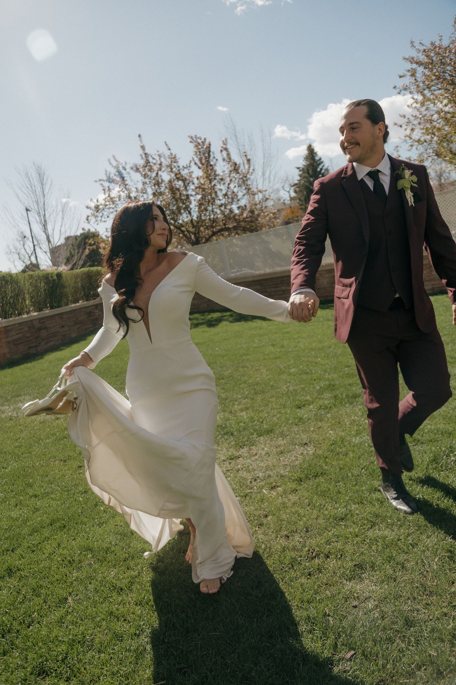 Bride in white gown and groom in burgundy suit hold hands running across a sunny green lawn.