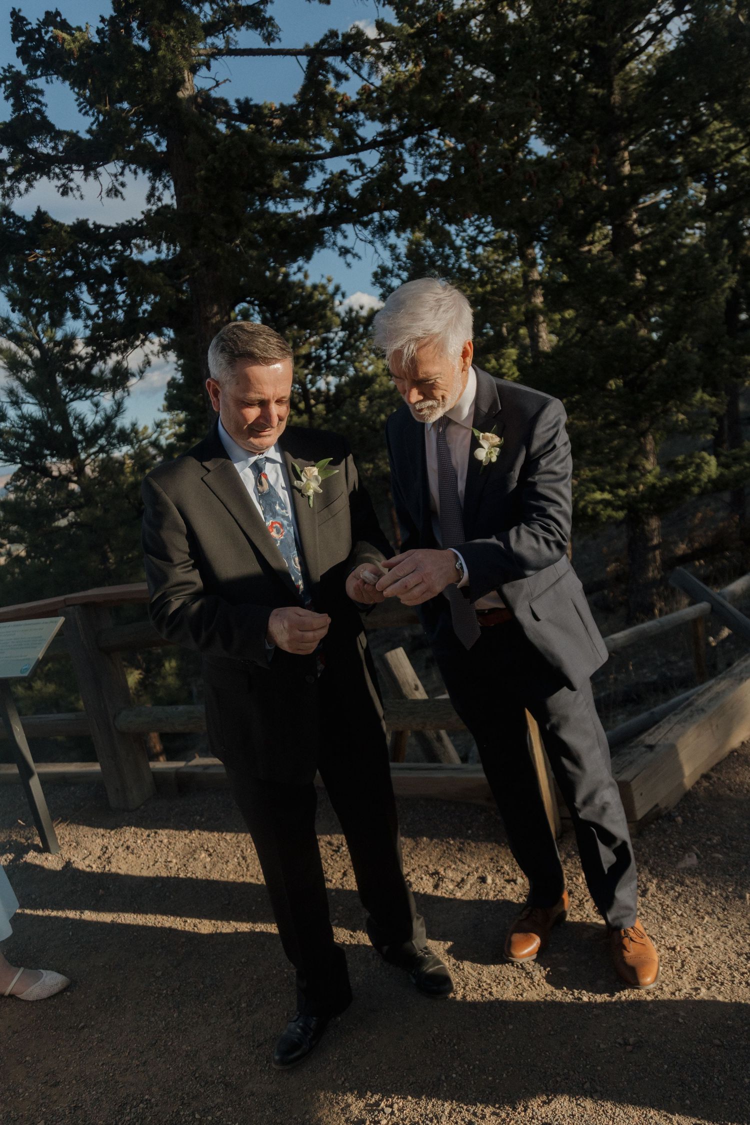 Two men in suits examining boutonniere outdoors at a wedding venue surrounded by pine trees.