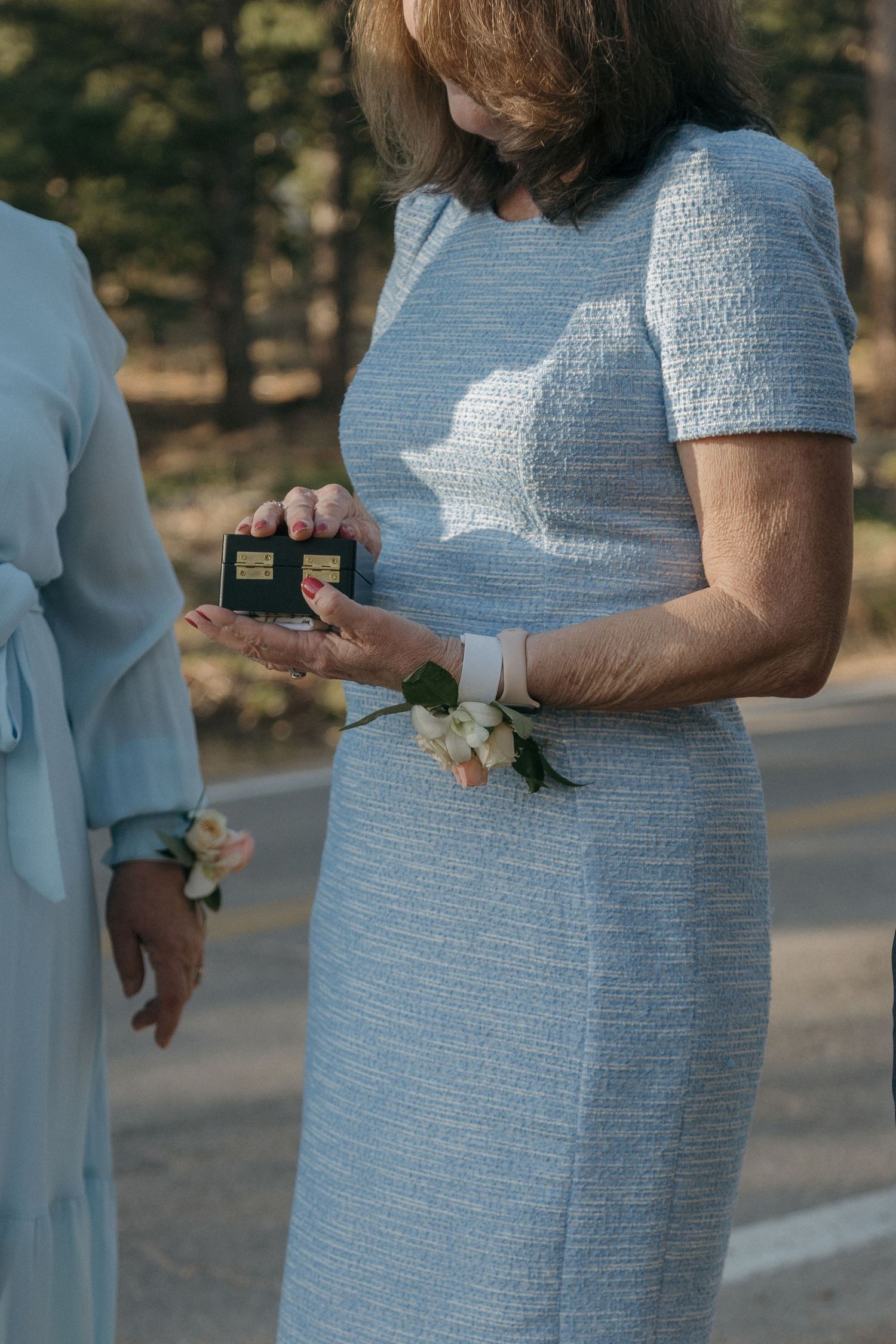 Woman in light blue tweed dress holding small ring box during outdoor wedding ceremony.