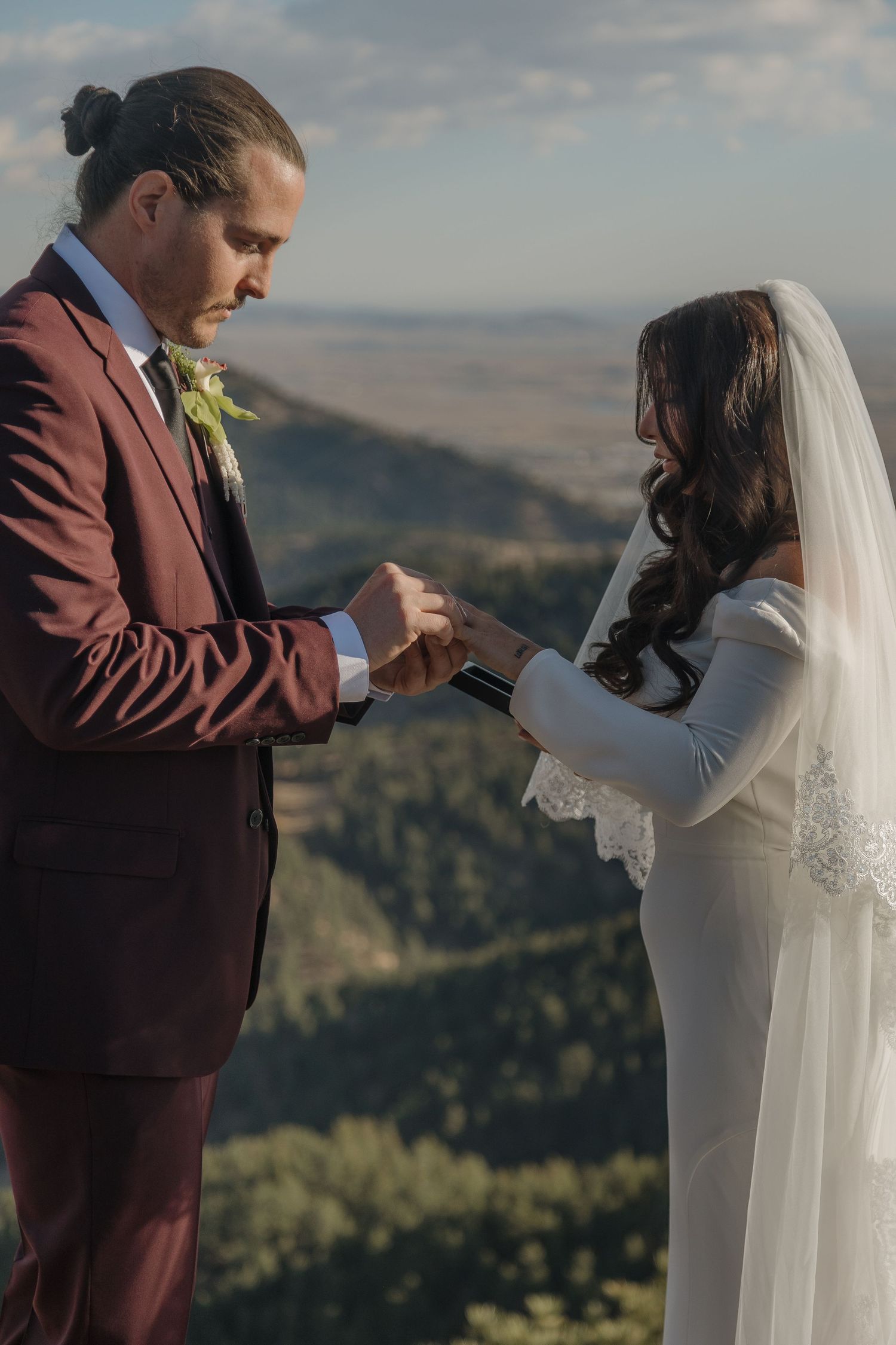 Groom in burgundy suit places ring on brides finger during outdoor mountain wedding ceremony.