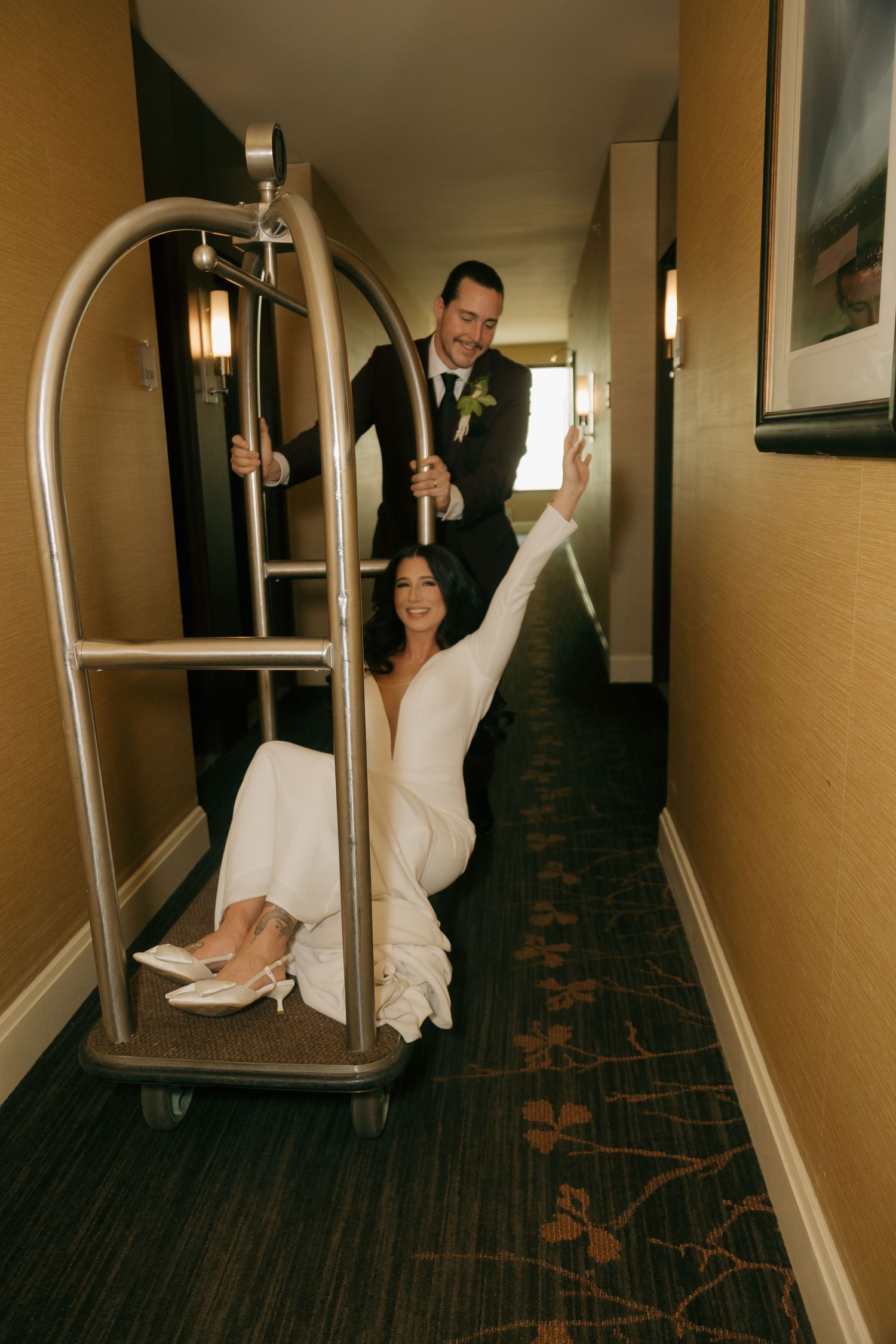 A playful couple poses with a hotel luggage cart in a hallway, creating a fun wedding photo moment.