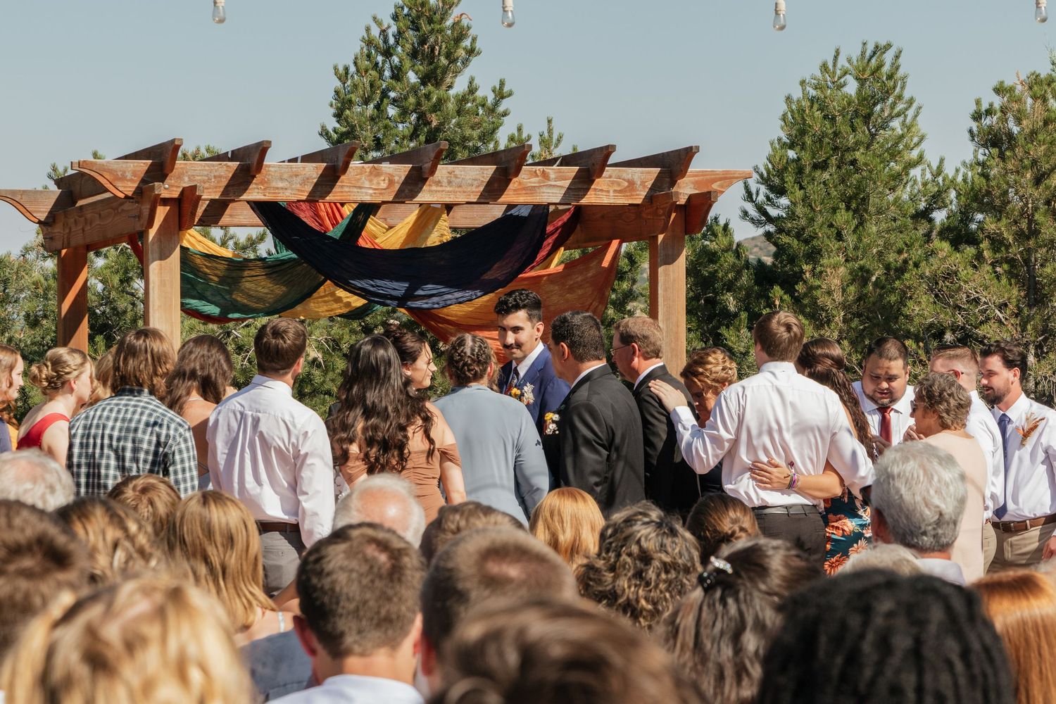 Guests gather under a wooden pergola for an outdoor wedding ceremony against a blue sky.