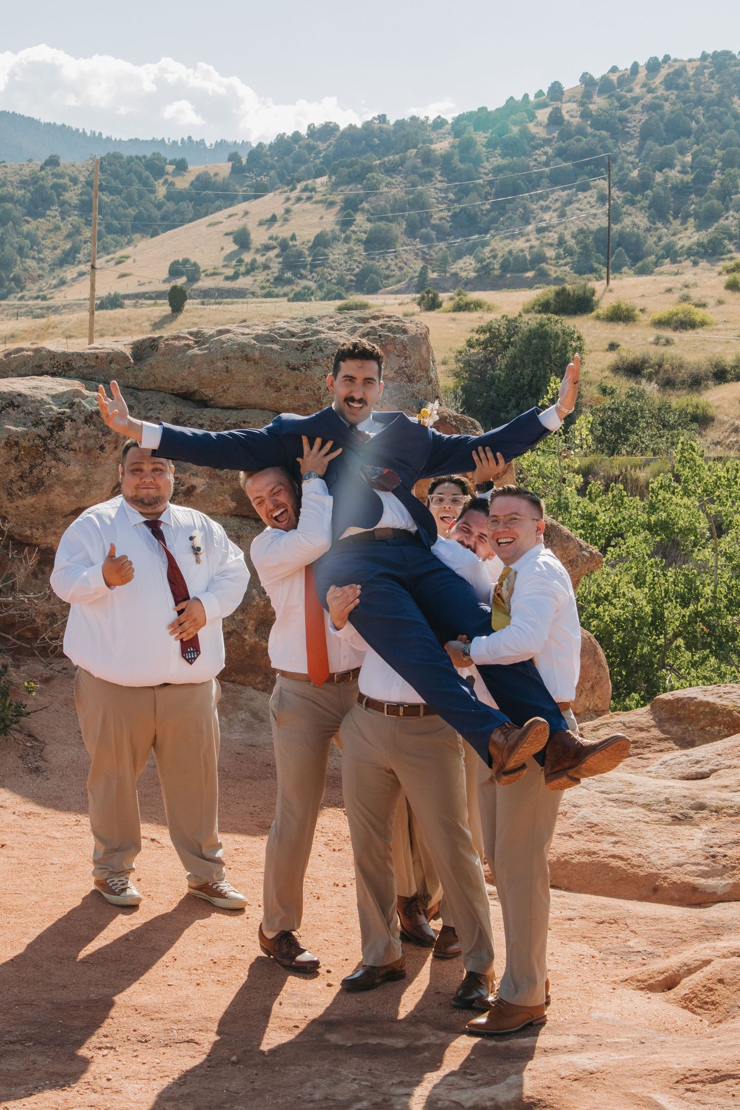 Groomsmen in khaki and navy suits playfully lifting and posing with groom against desert backdrop.