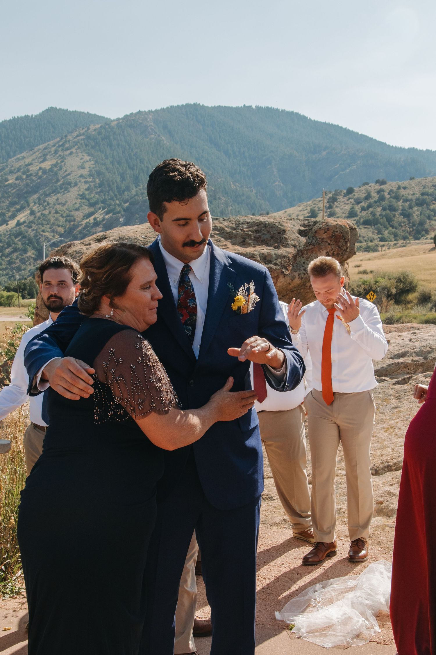 Joyful wedding party members embracing outdoors with mountains in background.