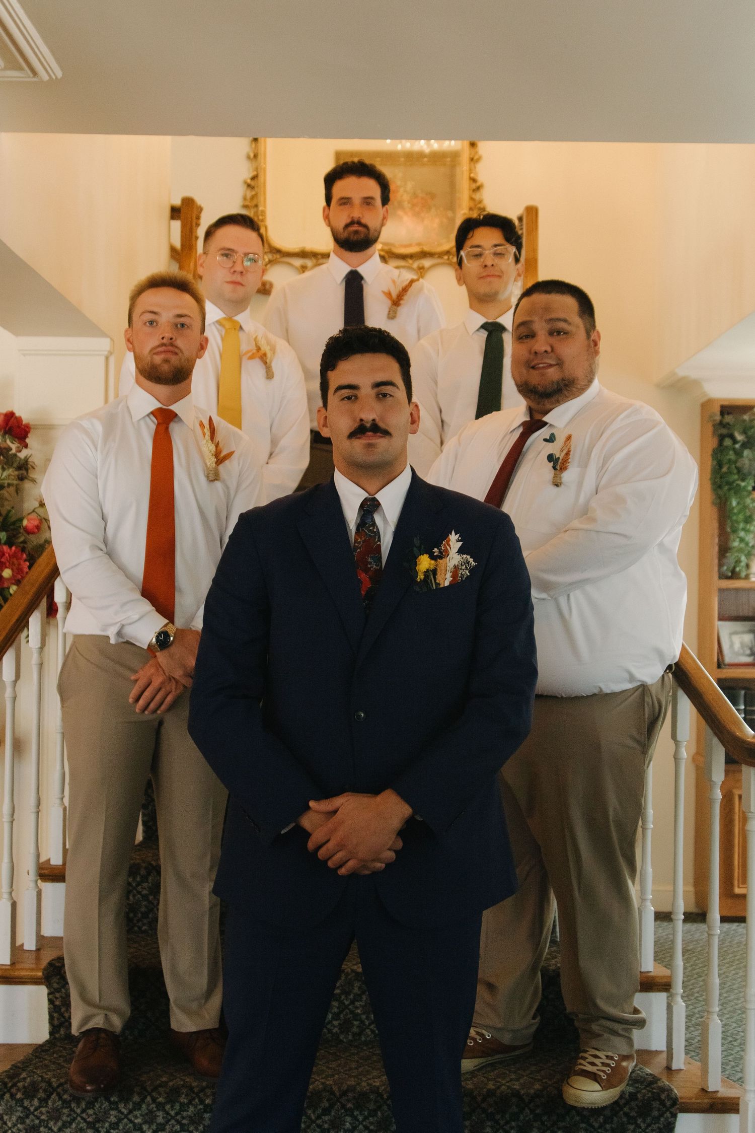 Wedding party members pose in formal attire on white stairs with a chandelier overhead.