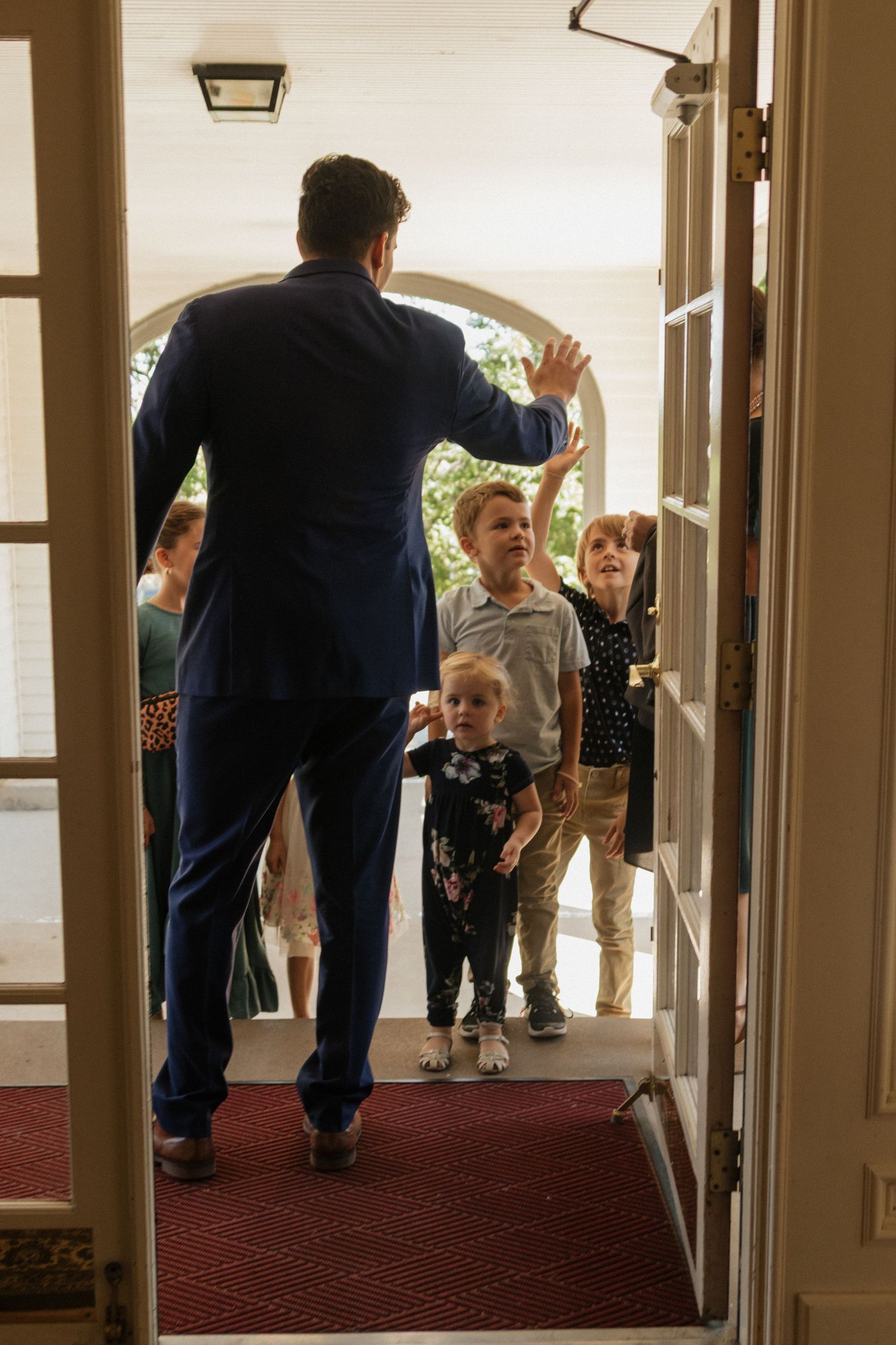Children and adults walk through an elegant hallway with wooden columns and red carpeting.