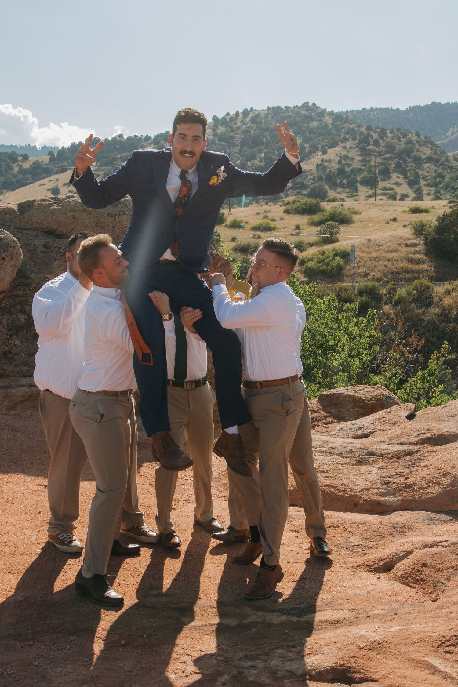 Groomsmen in white shirts and khakis pose playfully on red rocks with mountain views.