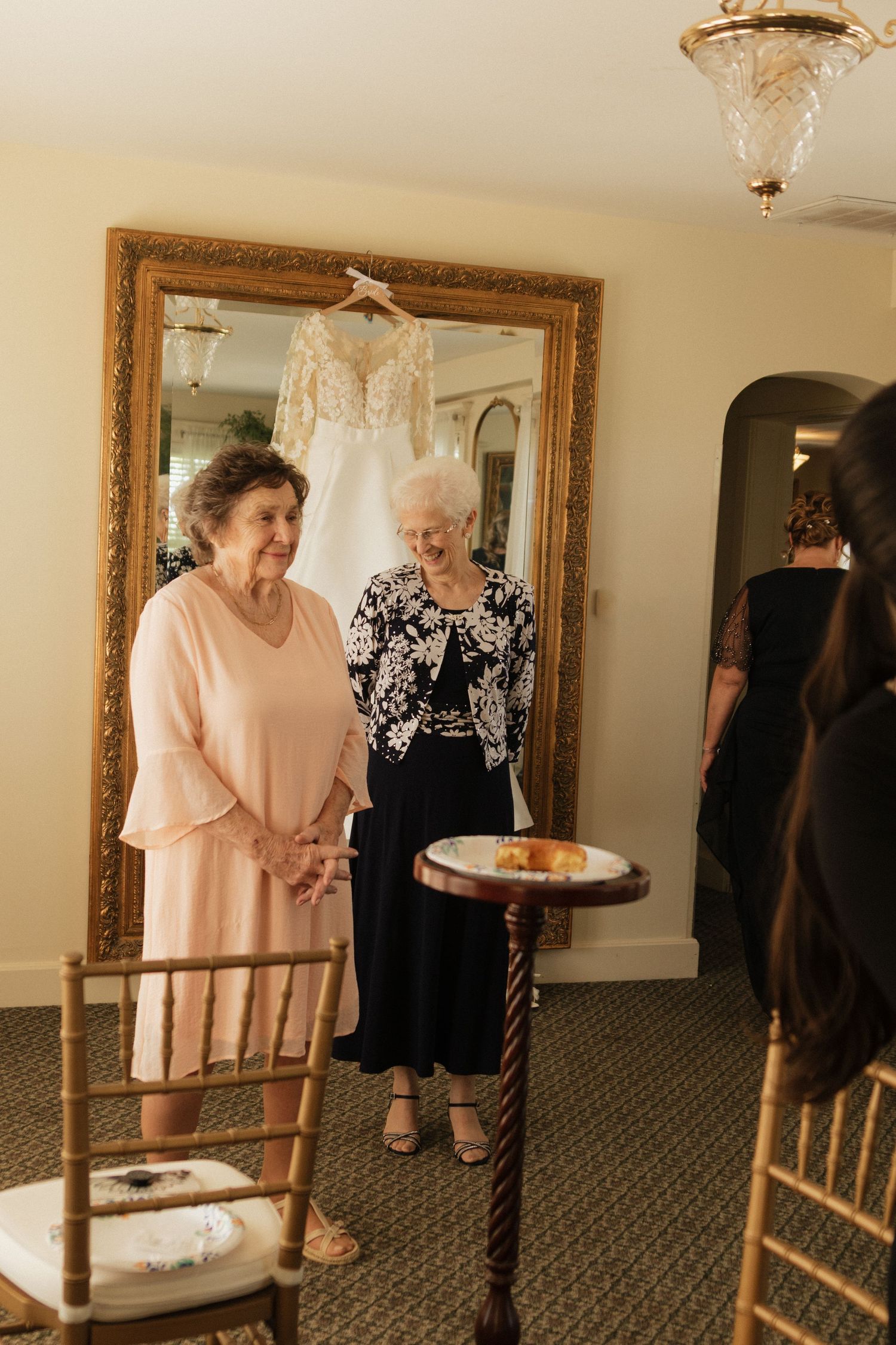 A wedding dress hangs on display in an ornate gold mirror while guests gather in the room.