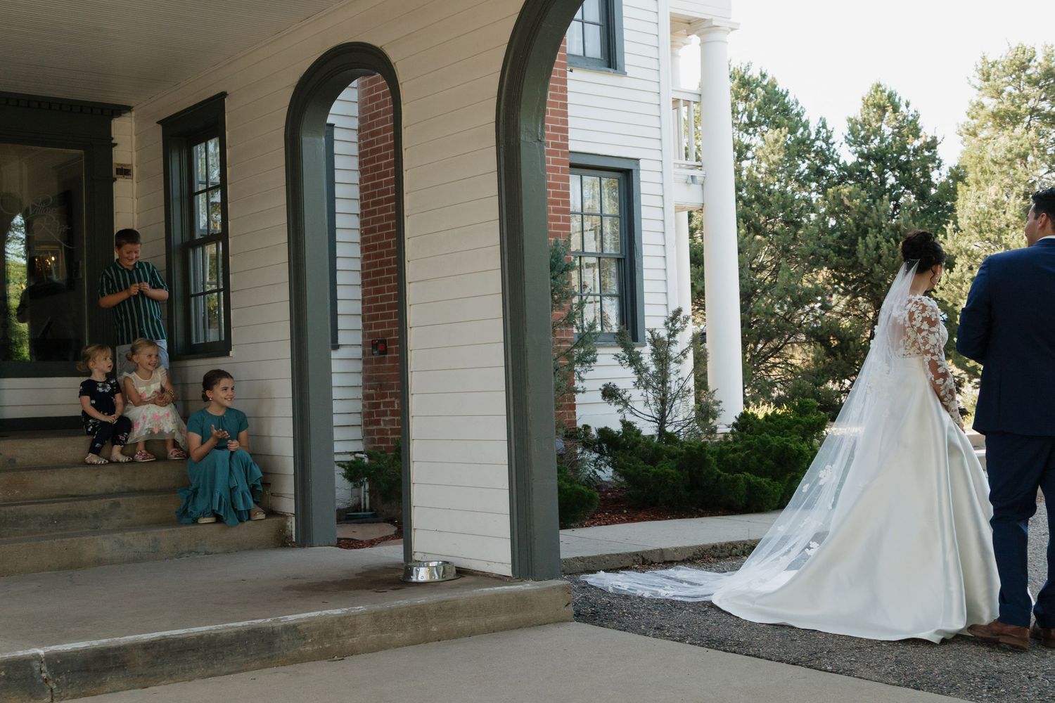 A flowing white wedding dress moves across the porch of a traditional white building.