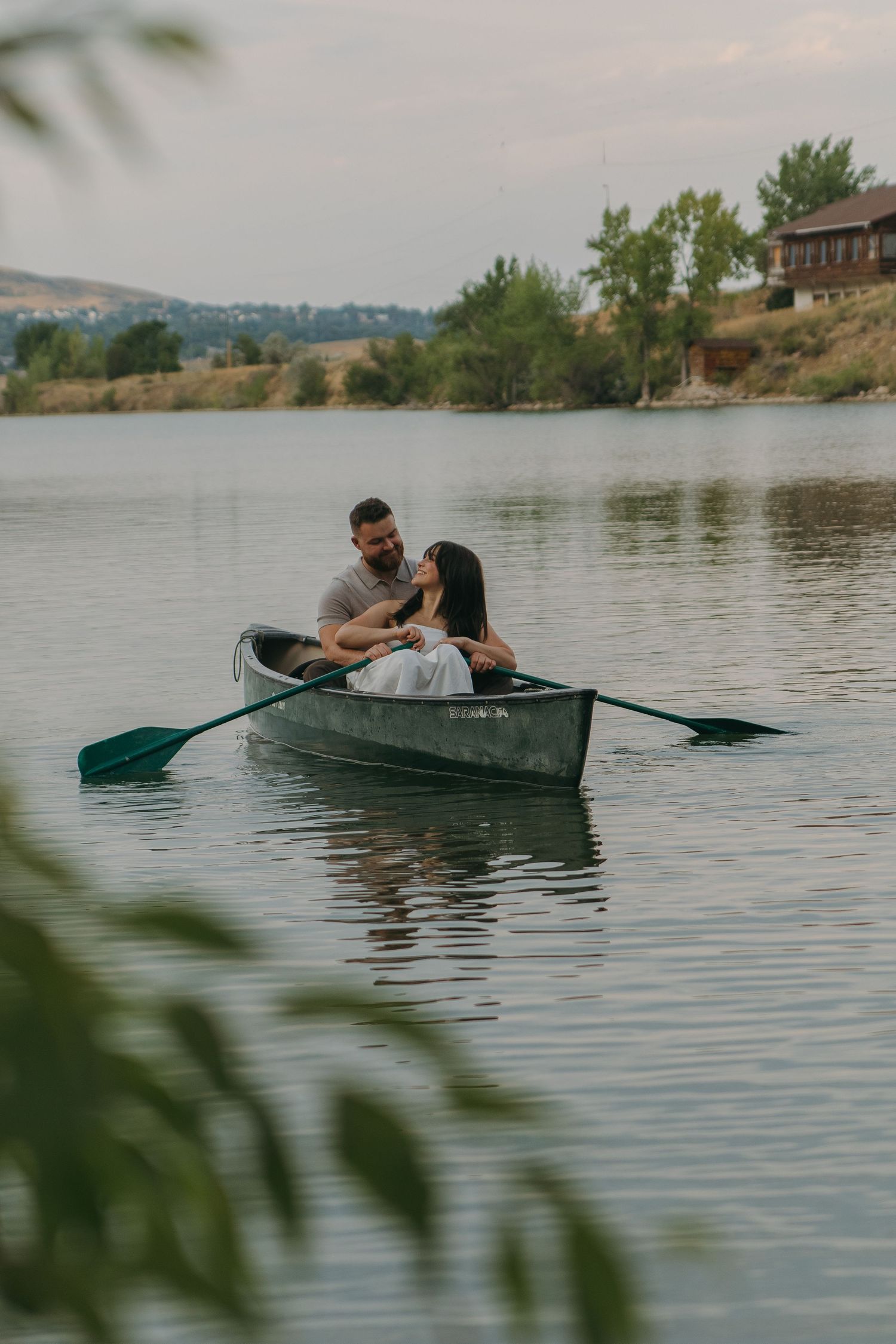 Two people paddle a dark green canoe across calm lake waters near a tree-lined shore.