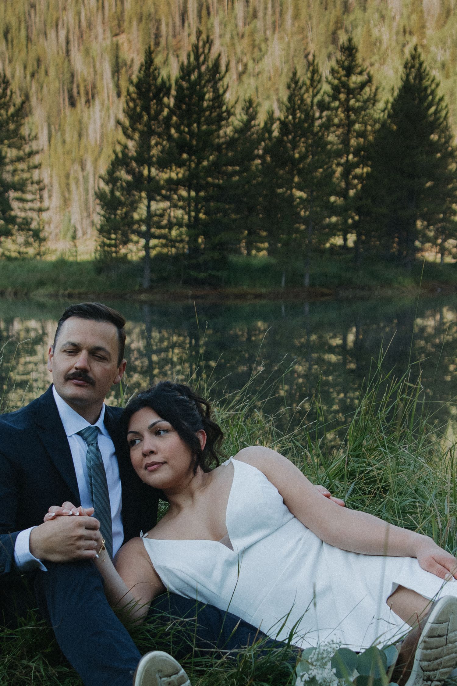An intimate wedding portrait by a mountain lake with pine trees reflecting in the water.