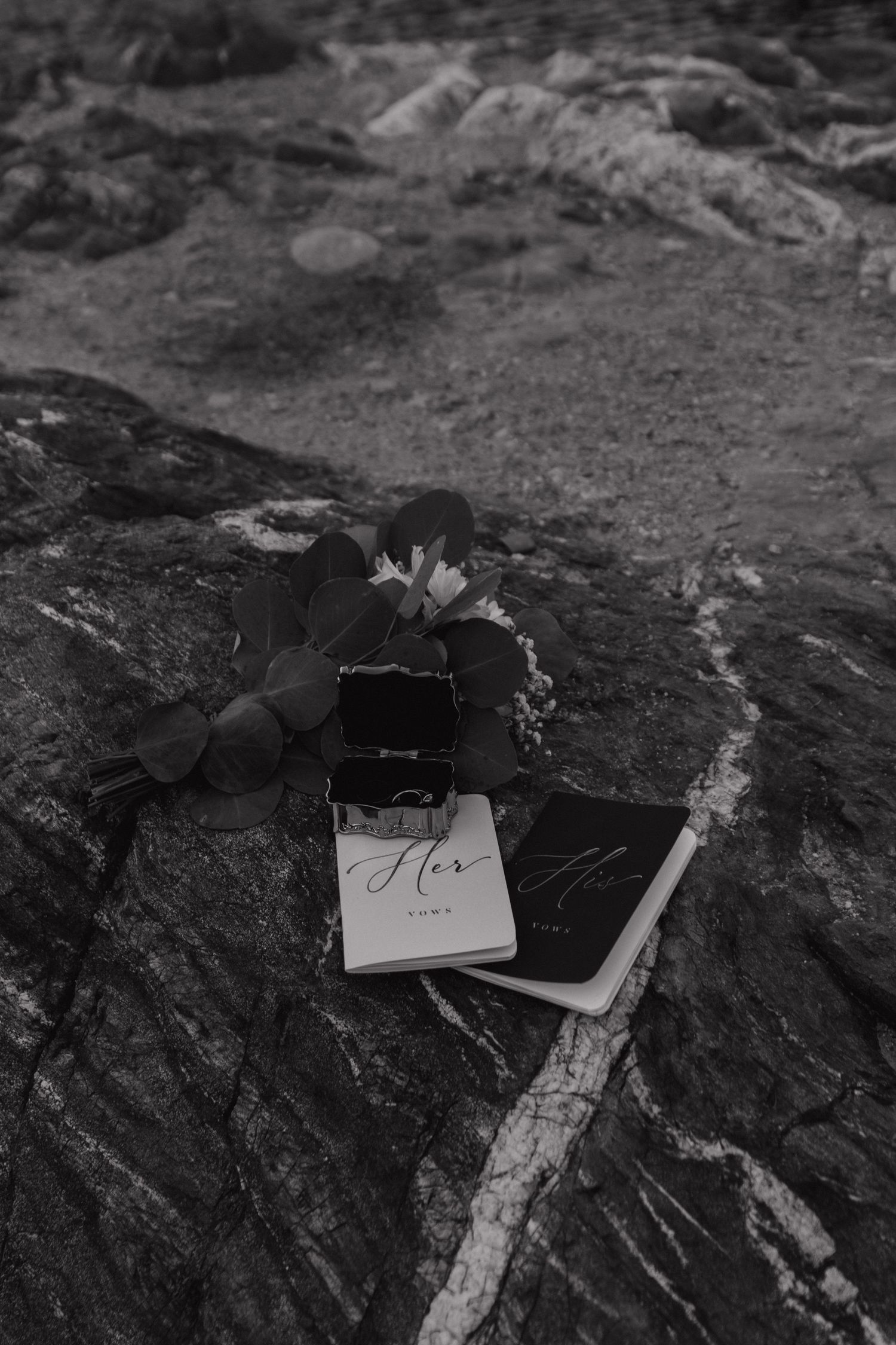Wedding vows and rings displayed on dark rocky surface in black and white.