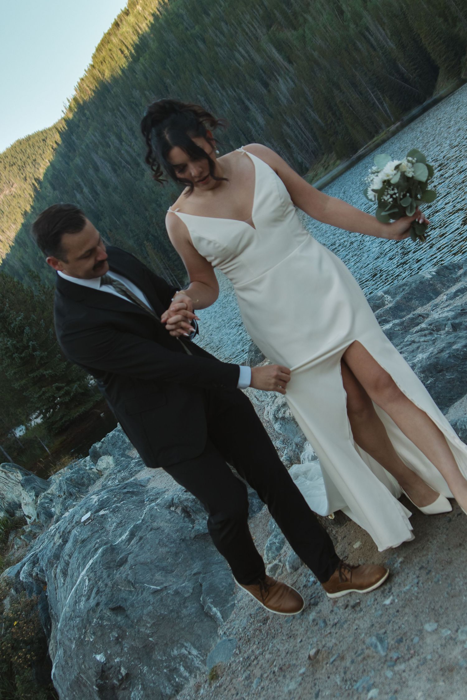 Newlyweds share a romantic moment on rocky terrain by a serene mountain lake.