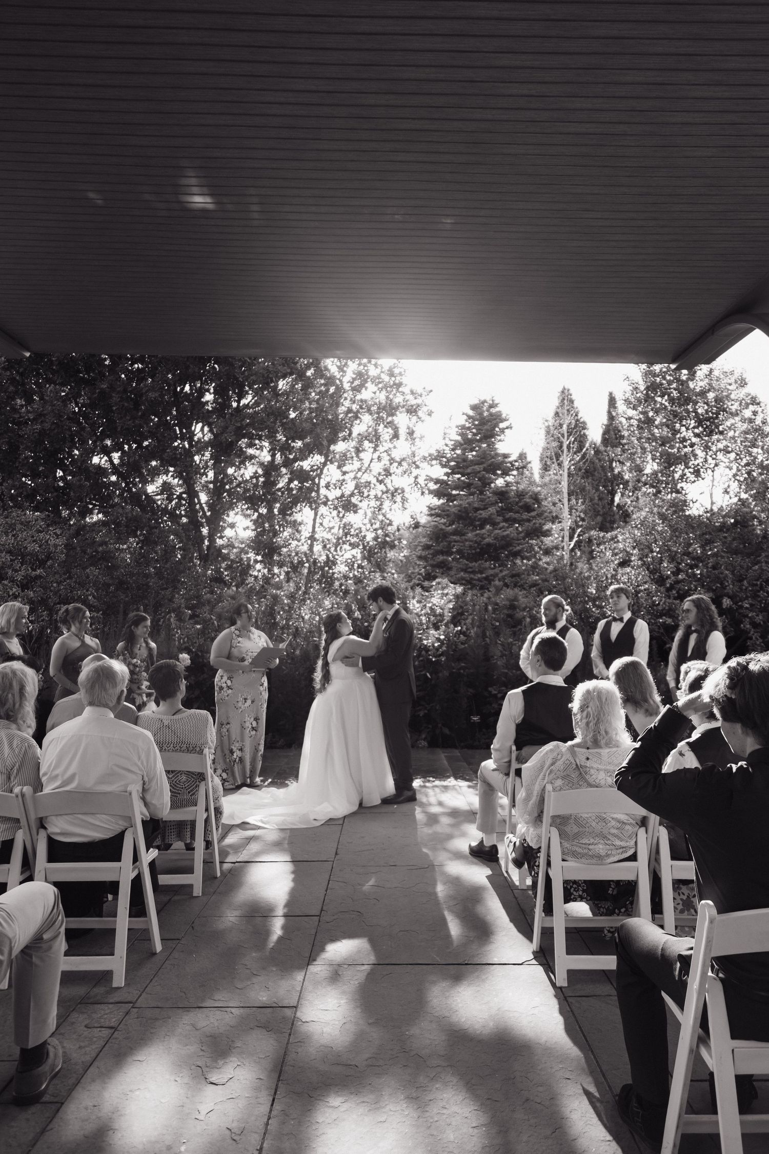 Black and white photo of a wedding ceremony with guests seated in white chairs as couple exchanges vows outdoors.