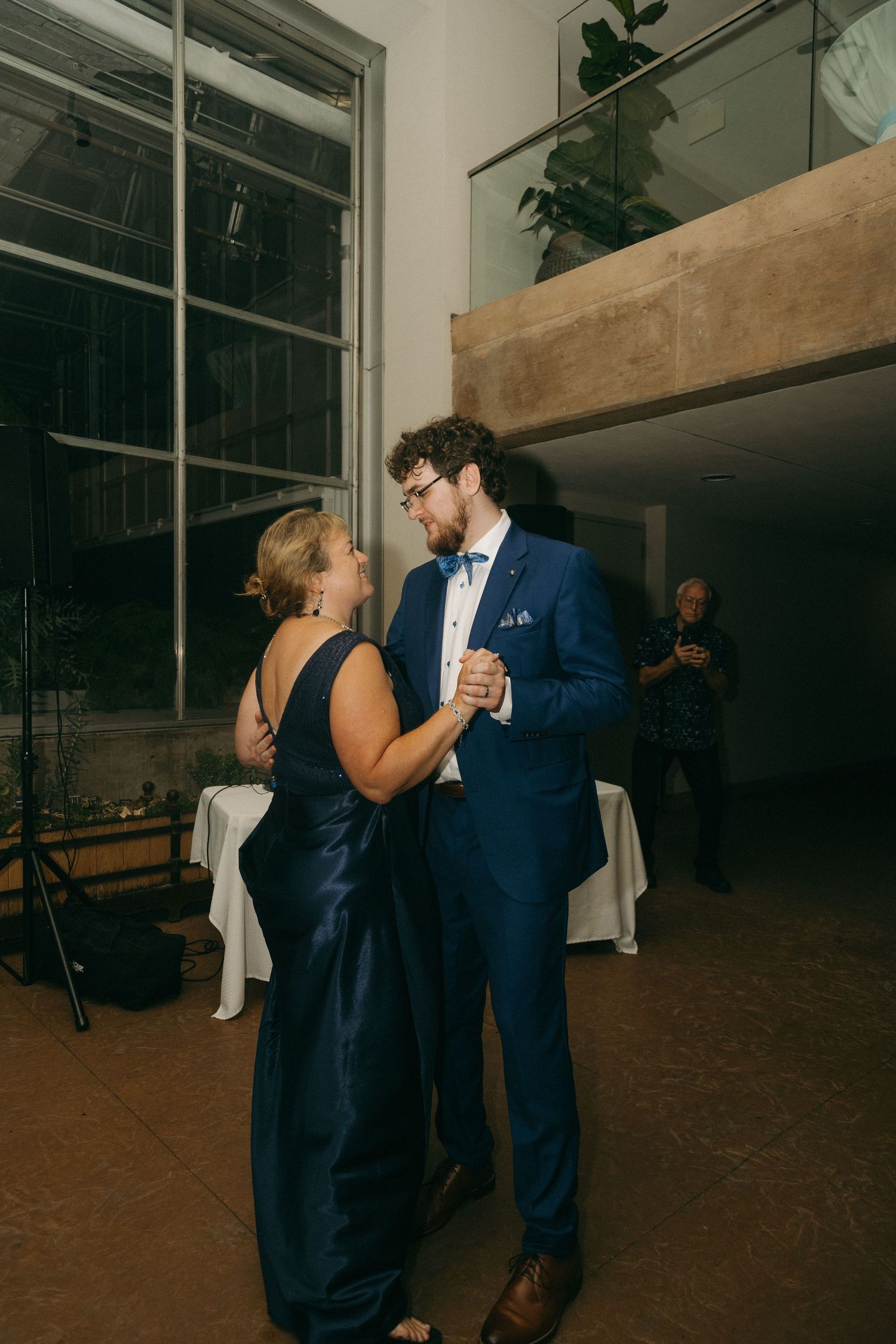 Groom in navy blue suit shares a mother-son dance at indoor wedding reception with large windows in background.
