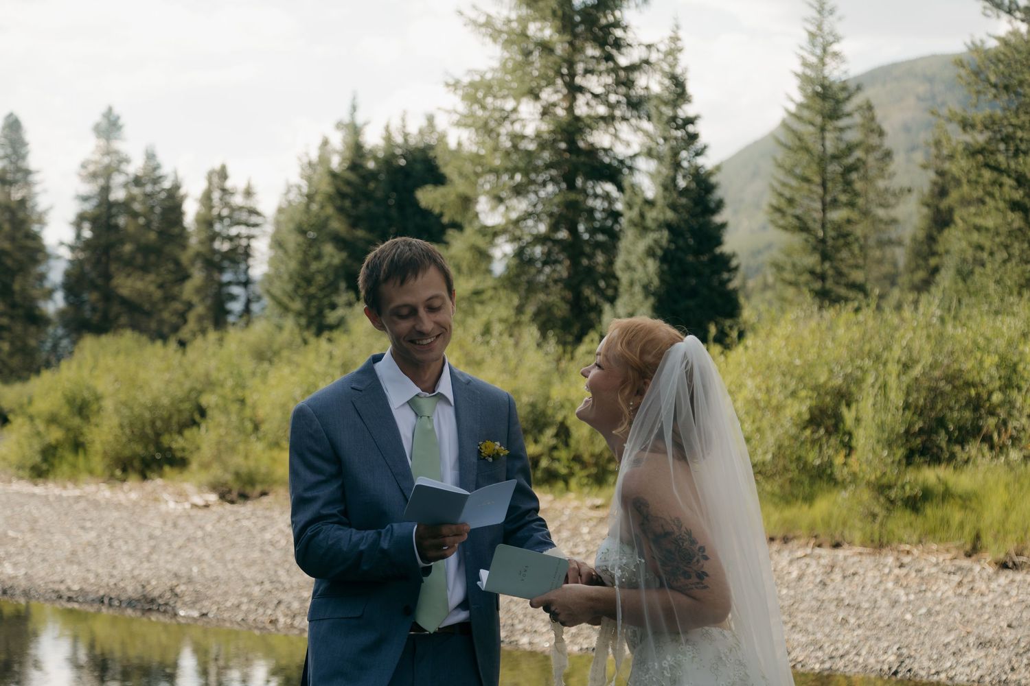 Groom in blue suit reads wedding vows to bride in white dress and veil outdoors near a mountain stream.