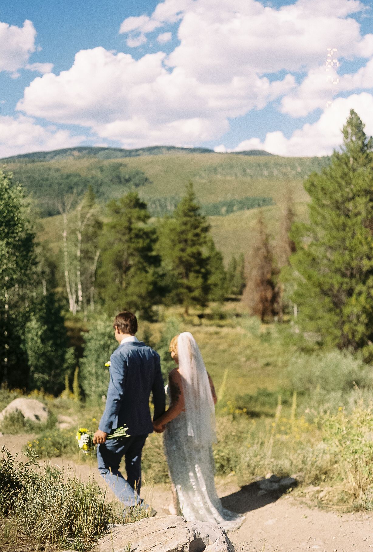 Bride and groom walk hand in hand on a mountain path surrounded by pine trees and a bright blue sky.