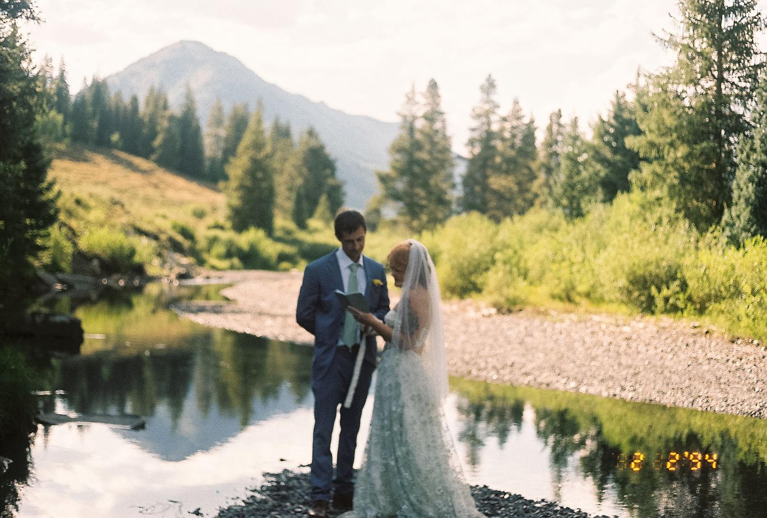 Bride and groom exchange vows beside a reflective mountain stream with pine trees and peaks in the background.