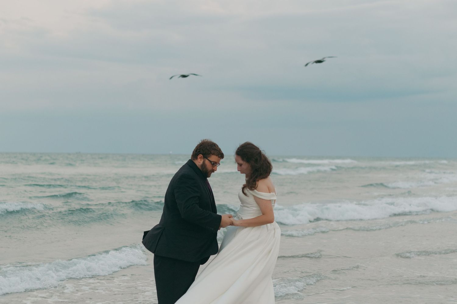 Couple in wedding attire holding hands on a beach with ocean waves and birds flying in the cloudy sky.