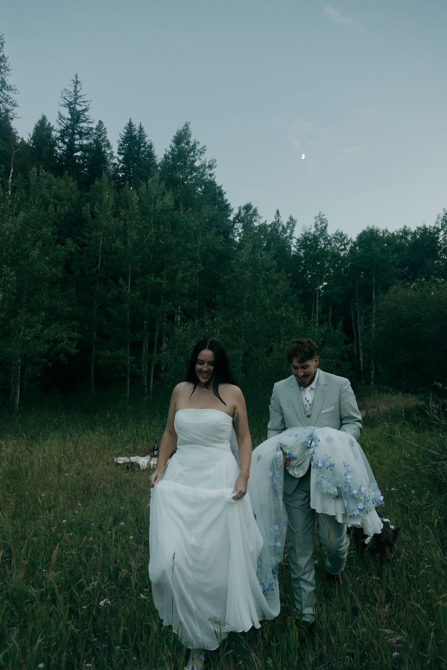 Bride and groom walking through a lush green forest meadow at dusk in wedding attire.