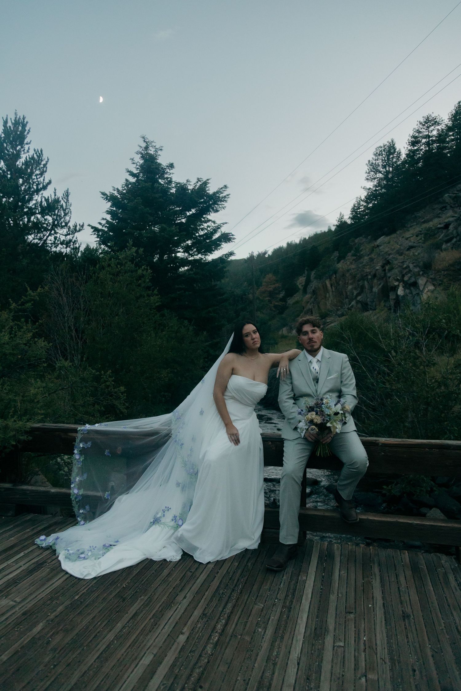 Bride in flowing white gown with veil and groom in gray suit posing on wooden deck surrounded by forest at dusk.