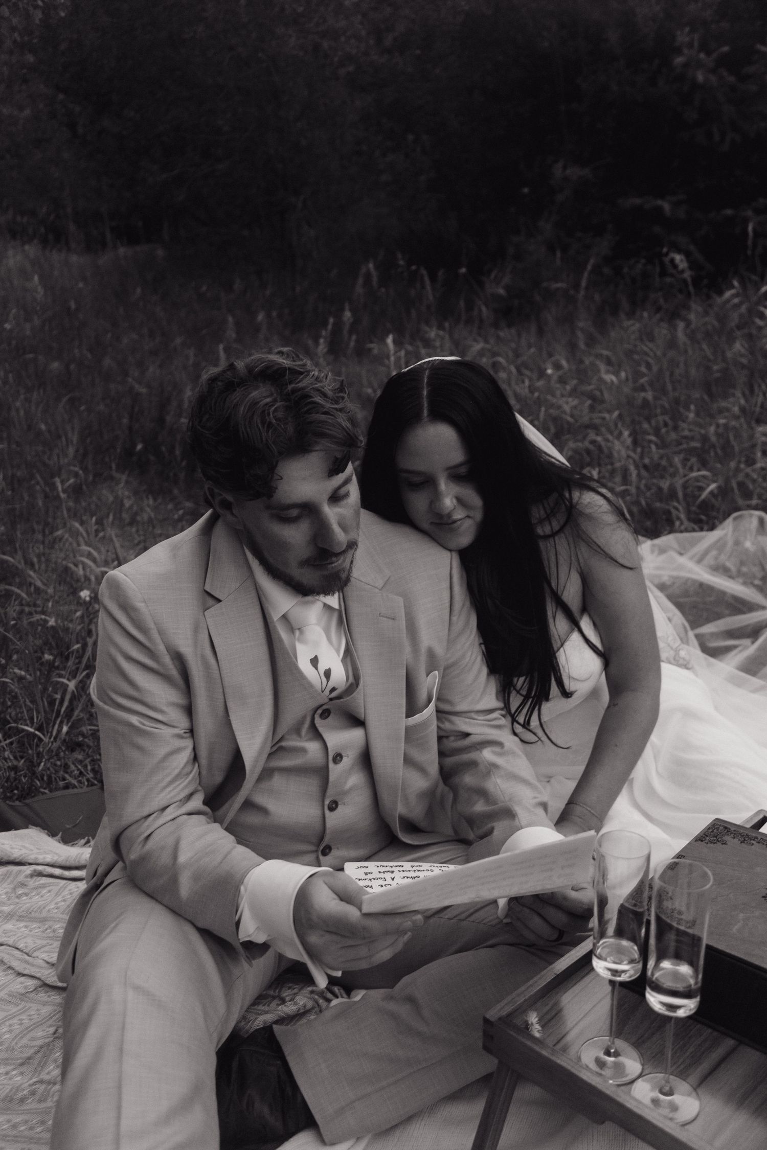 Black and white photo of bride and groom reading vows on a blanket in a field with champagne glasses nearby.