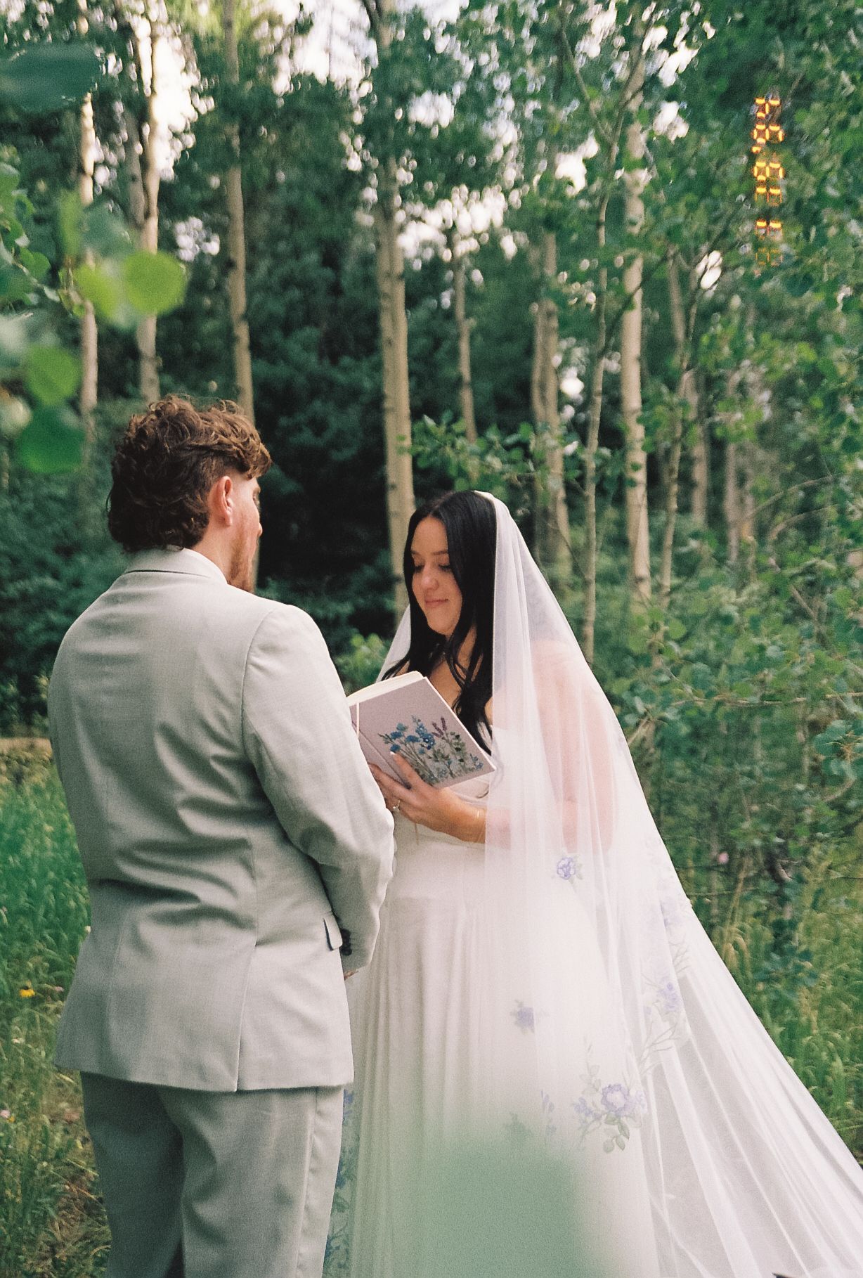 Bride in white gown and long veil reads wedding vows from a book to her groom among tall aspen trees in a forest ceremony.