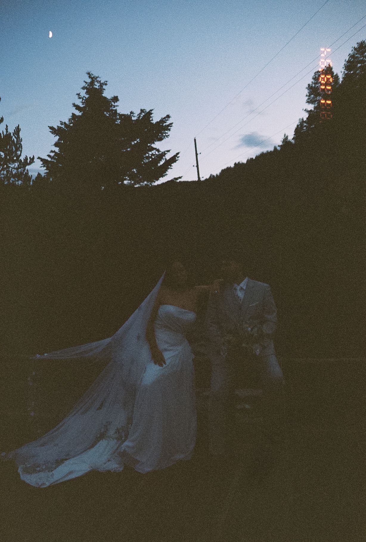 Silhouette of bride in flowing veil and groom embracing under twilight sky with trees and lights in background.