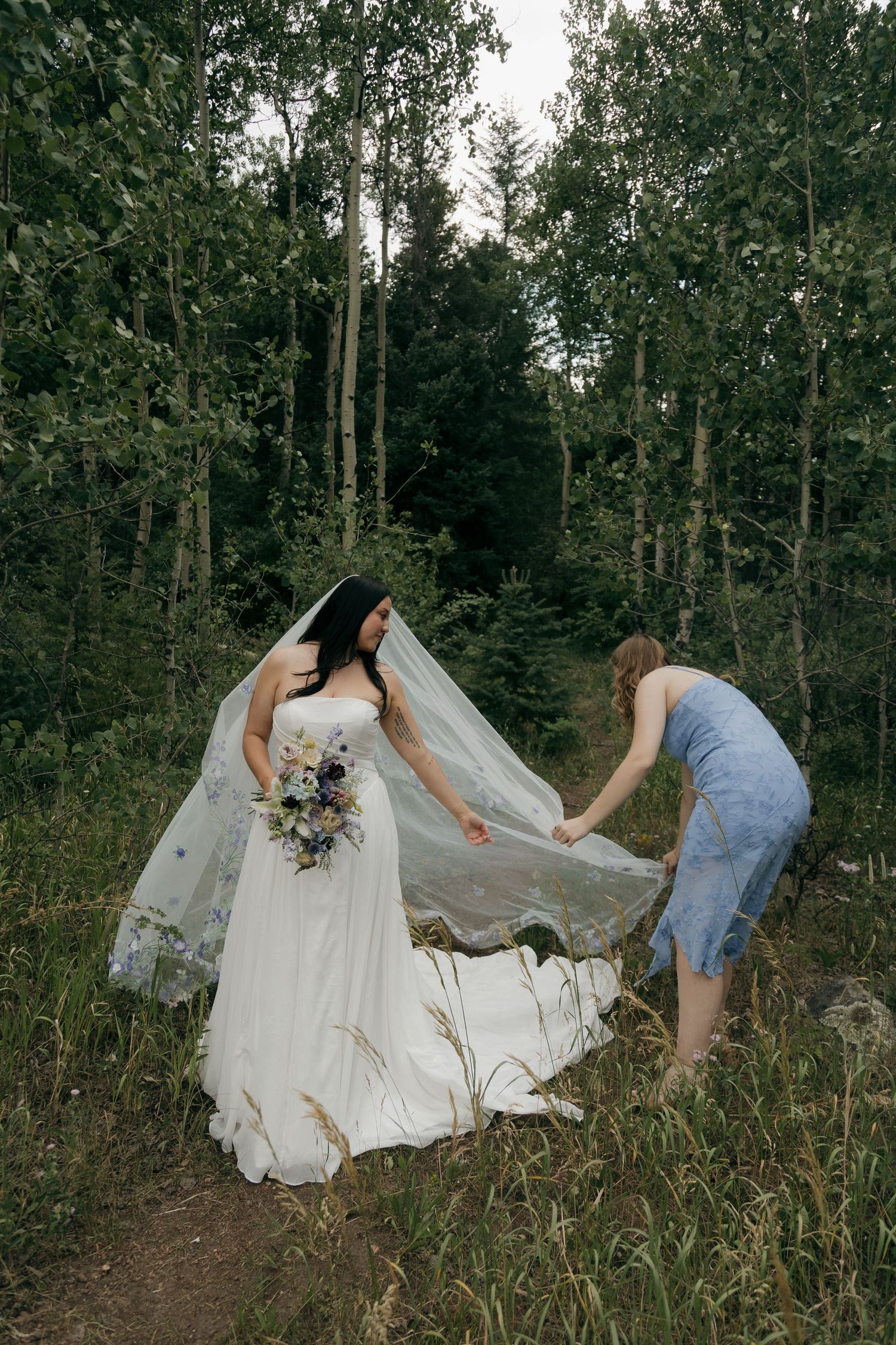 Bridesmaid adjusting bride's floral veil in a lush forest setting, bride holding a wildflower bouquet.