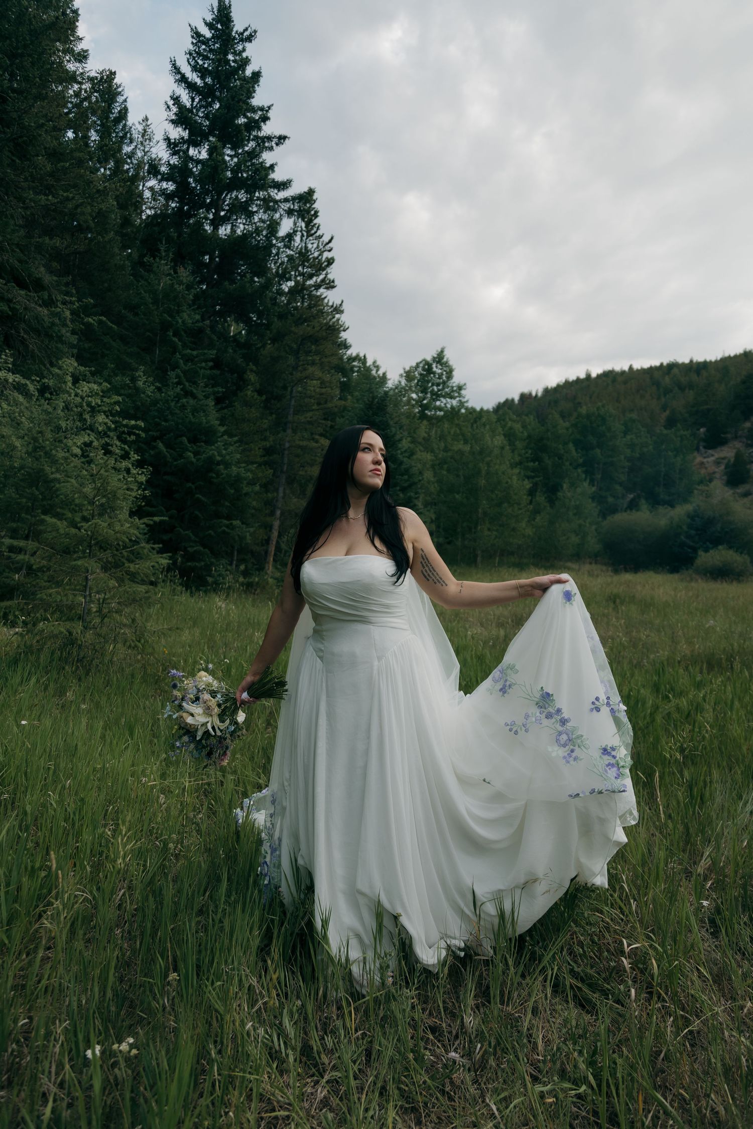 Bride in white embroidered gown holding wildflower bouquet in lush green mountain meadow with evergreen forest backdrop.
