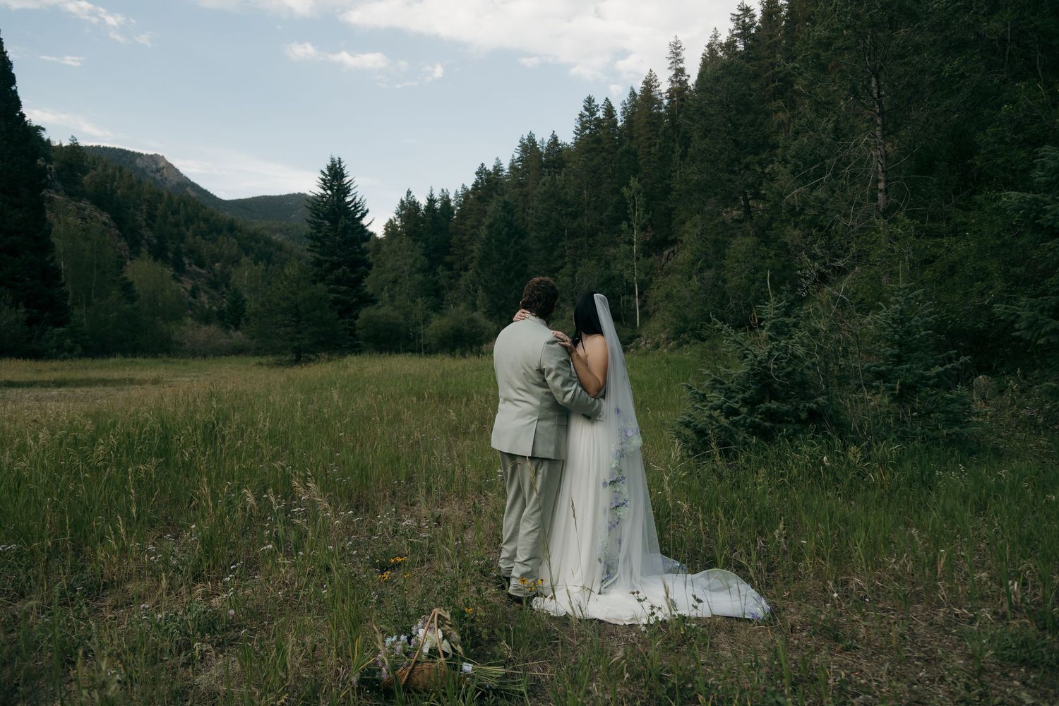 Bride and groom stand together gazing at scenic mountain meadow after outdoor elopement.