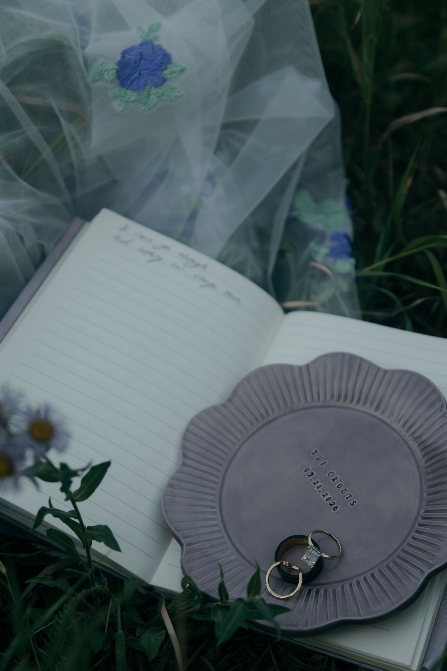 Wedding rings rest on gray dish atop open vow journal beside floral embroidered veil in grass.