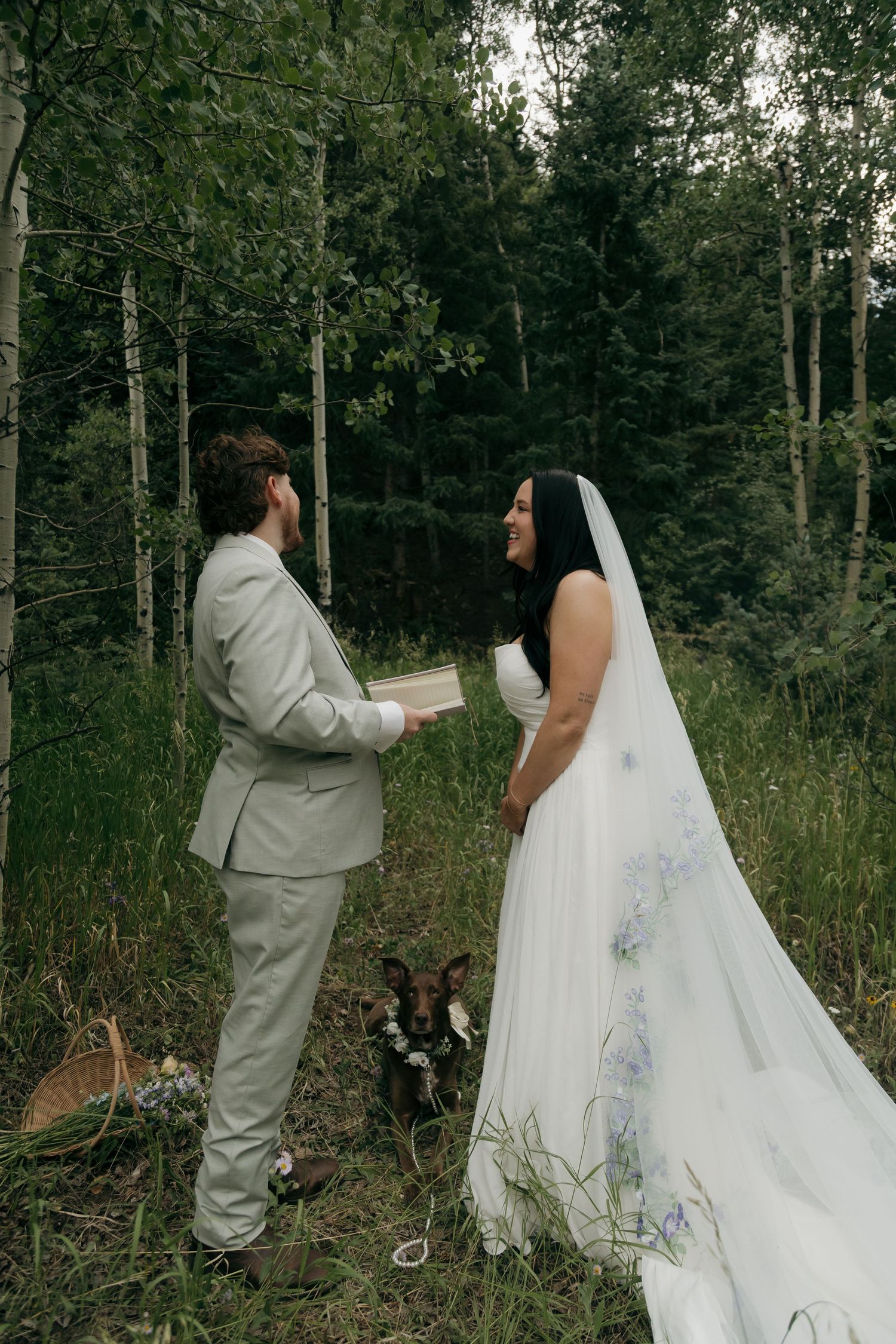 Bride and groom exchange vows in a lush forest setting, with a dog sitting nearby during their intimate woodland wedding ceremony.
