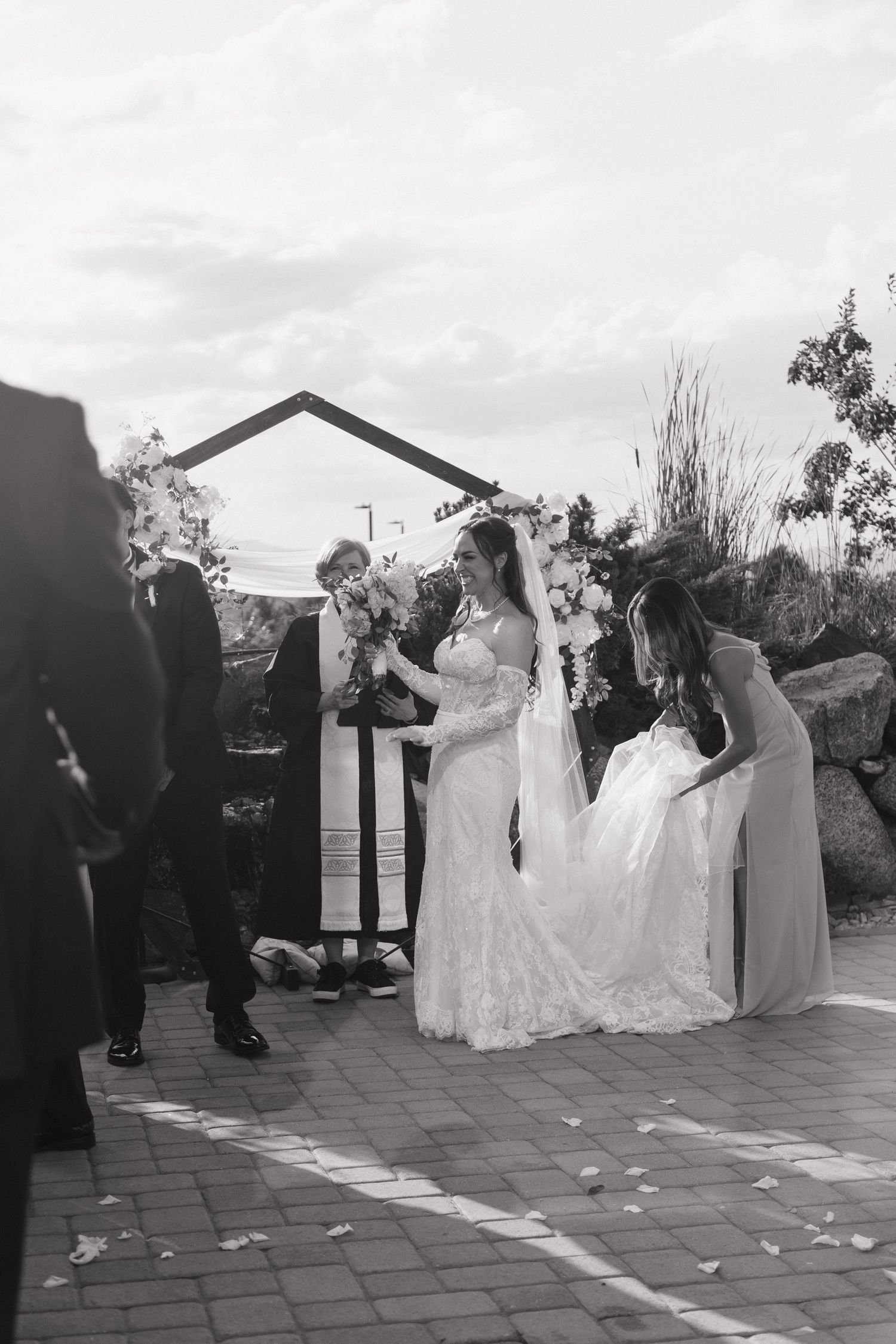 Bride in lace gown stands at outdoor altar with groom and officiant as bridesmaid adjusts her dress train.