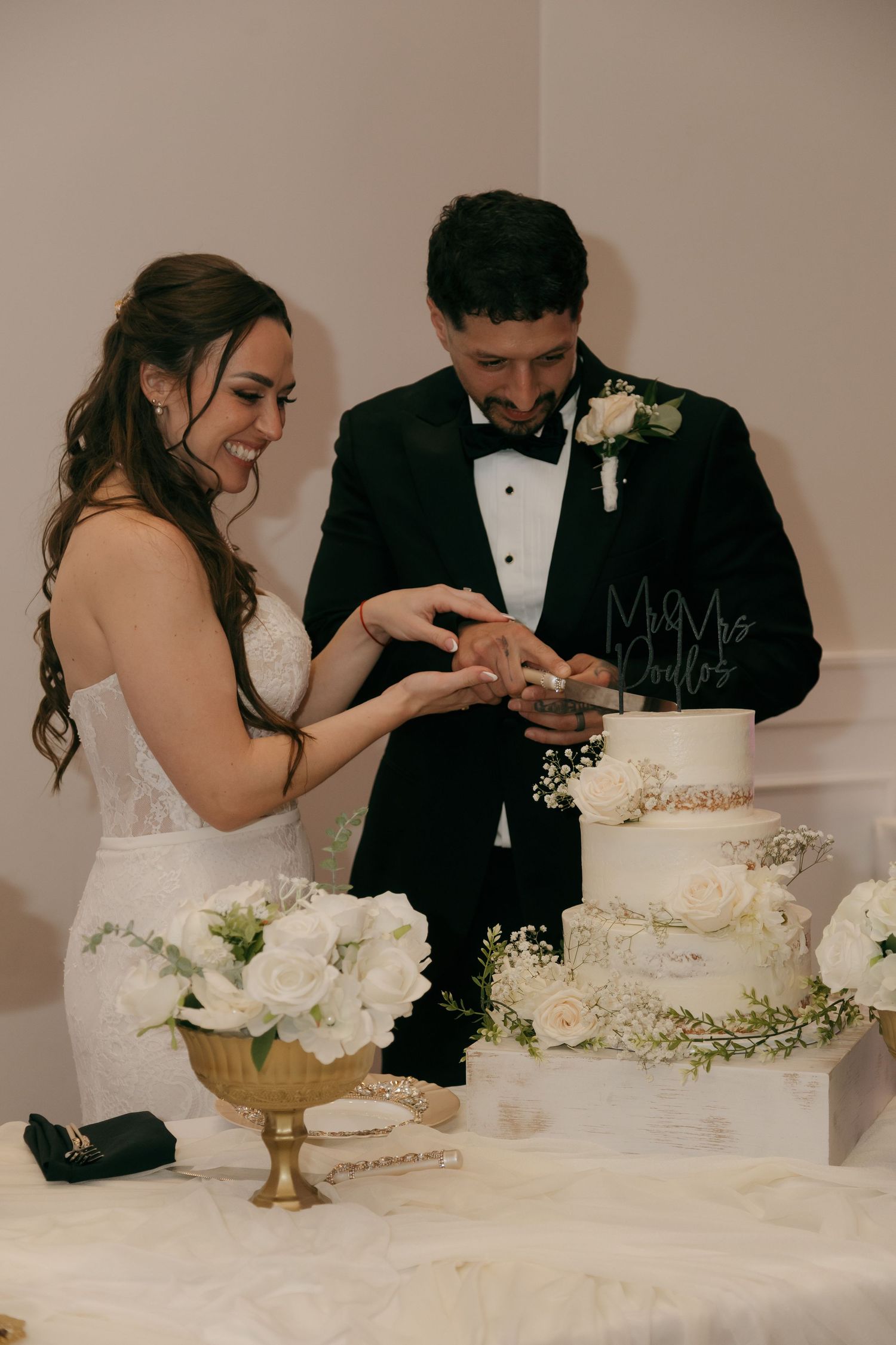 Smiling bride and groom in tuxedo cutting three-tier white wedding cake with floral decorations.