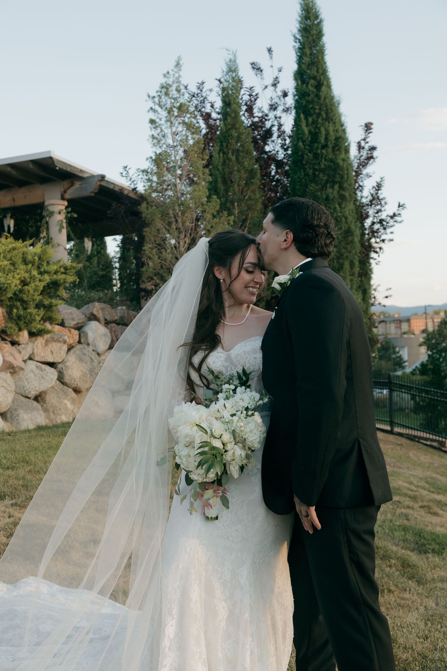 Groom in black suit kisses bride in white lace gown holding bouquet at outdoor wedding venue.